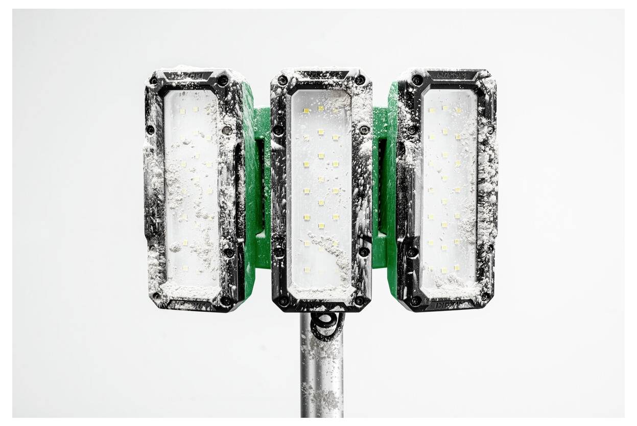 Trzy prostokątne przemysłowe lampy LED pokryte kurzem, zamocowane na metalowym słupie, na białym, jednolitym tle.