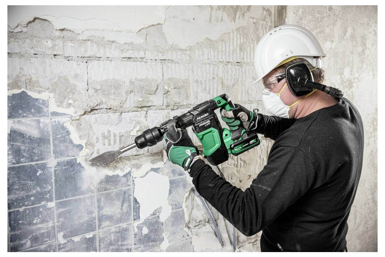 Osoba w odzieży ochronnej używa zielonego elektronarzędzia do usuwania starych płytek ze ściany, nosząc kask, okulary ochronne i maskę dla własnego bezpieczeństwa.