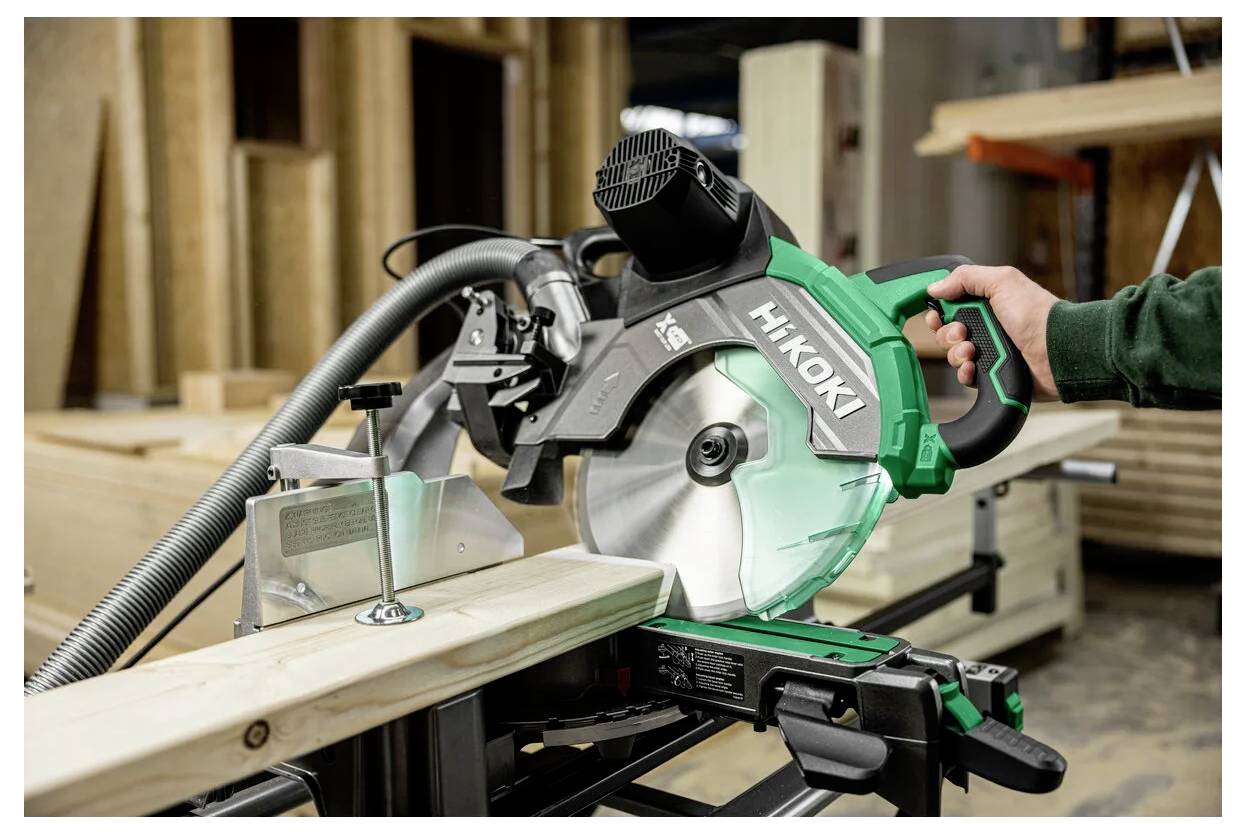 Osoba obsługuje zieloną piłę ukosową Hikoki, przecinająca drewnianą deskę w warsztacie, z różnymi materiałami drewnianymi w tle.