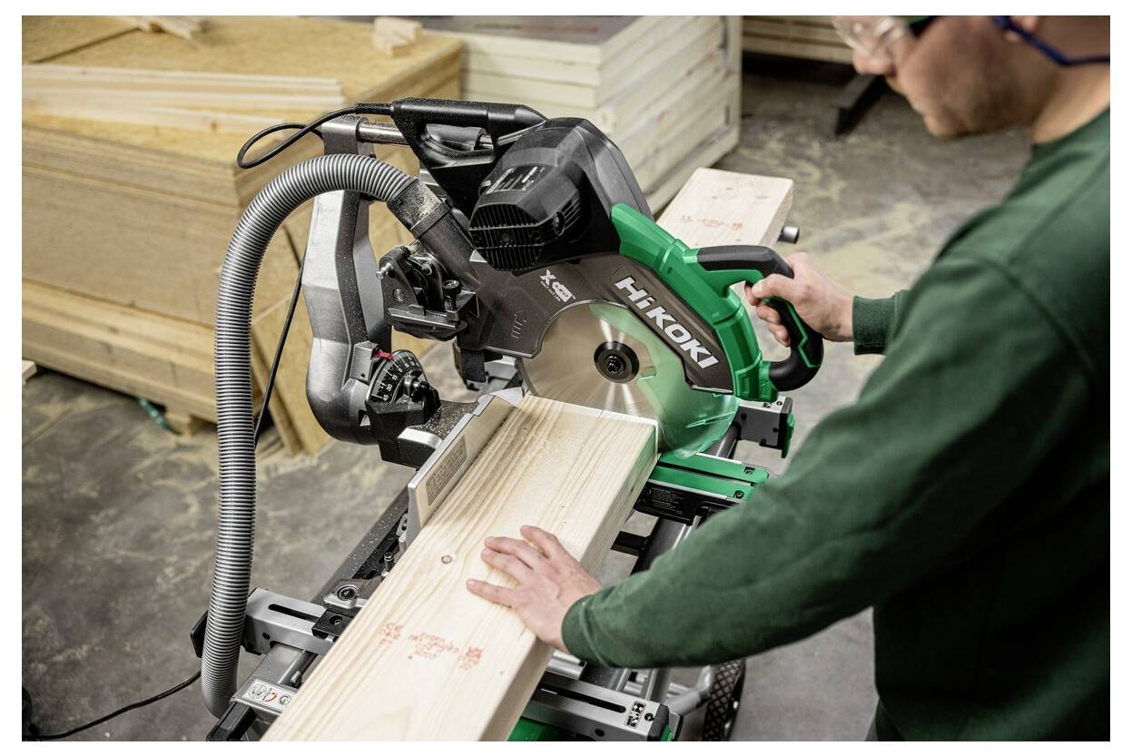 Osoba używająca zielonej i czarnej piły ukośnej Hitachi do cięcia deski drewnianej na stole warsztatowym w warsztacie.