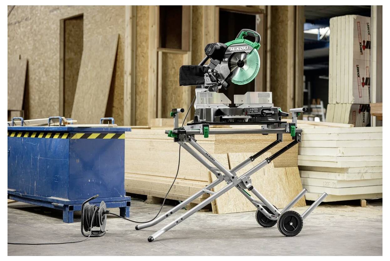Piła ukośnica na kółkach w warsztacie, z deskami drewnymi i niebieskim pojemnikiem na odpady w tle, sugerujące prace budowlane.