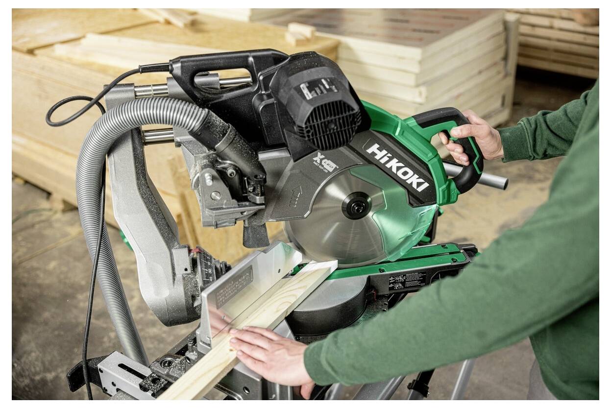 Osoba w zielonej koszulce używa piły ukosowej do cięcia kawałka drewna w warsztacie. Piła jest oznaczona marką 'Hikoki'.