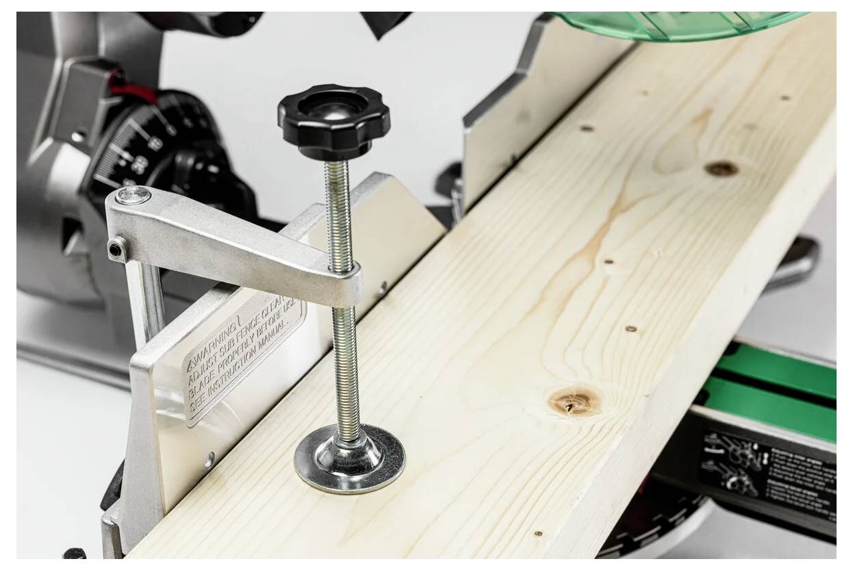 Drewniana deska jest mocno zaciśnięta w pilarce ukosowej z czarną dźwignią regulacyjną, przygotowana do cięcia. Widoczna jest skala pilarki.