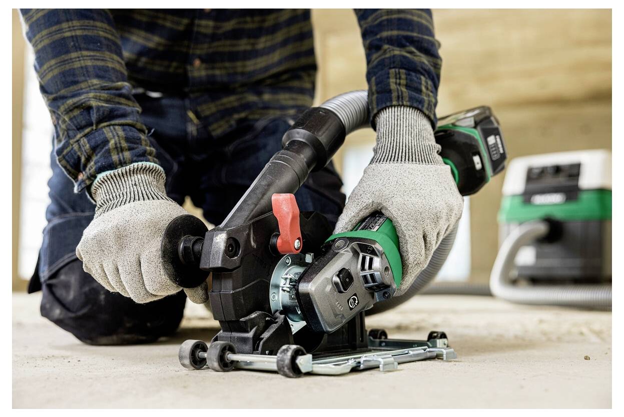 Osoba klęczy podczas używania ręcznego narzędzia elektrycznego do cięcia powierzchni, nosząc rękawice ochronne. Do urządzenia podłączony jest system odkurzania w celu kontroli pyłu.