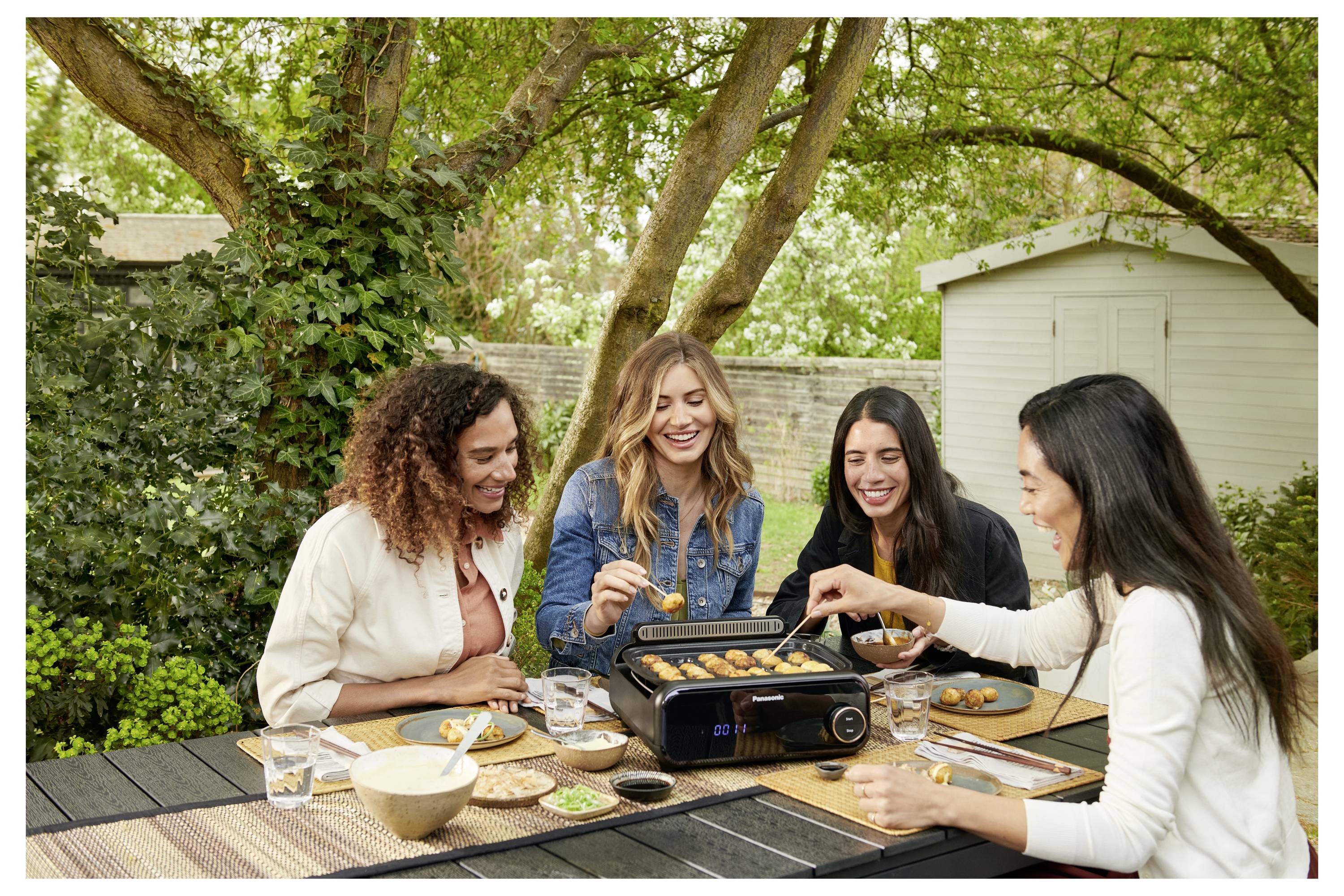 Cztery kobiety cieszą się gotowaniem i jedzeniem przy stole na wolnym powietrzu, otoczonym zielenią. Na stole umieszczony jest grill.