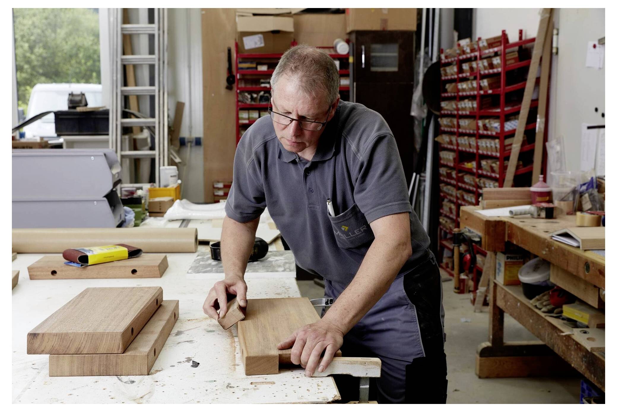 Mężczyzna pracuje w warsztacie, szlifuje drewnianą deskę na stole. W tle narzędzia i materiały są poustawiane na półkach i blatach.