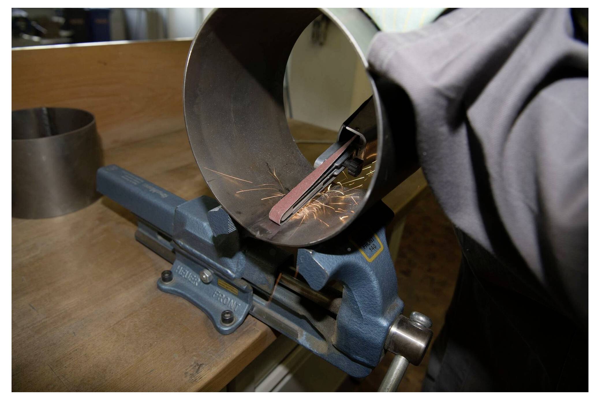 Pracownik używa pilnika wewnątrz metalowego cylindra zamocowanego w imadle, powodując powstawanie iskier. Otoczenie przypomina warsztat lub środowisko przemysłowe.