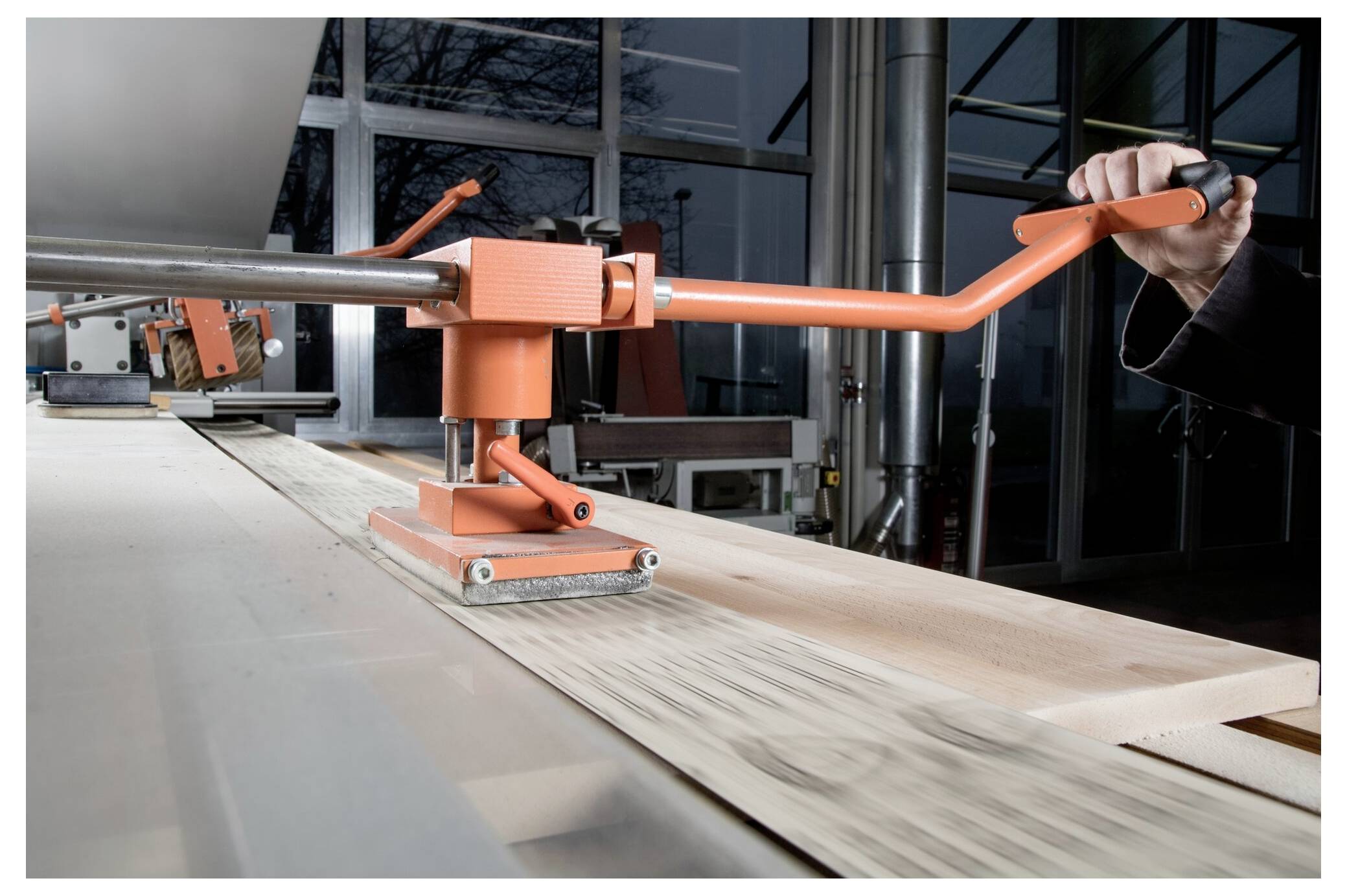 Osoba obsługuje przemysłowy uchwyt ręczny z pomarańczową rączką, mocując drewnianą deskę na metalowym stole warsztatowym w środowisku fabrycznym.