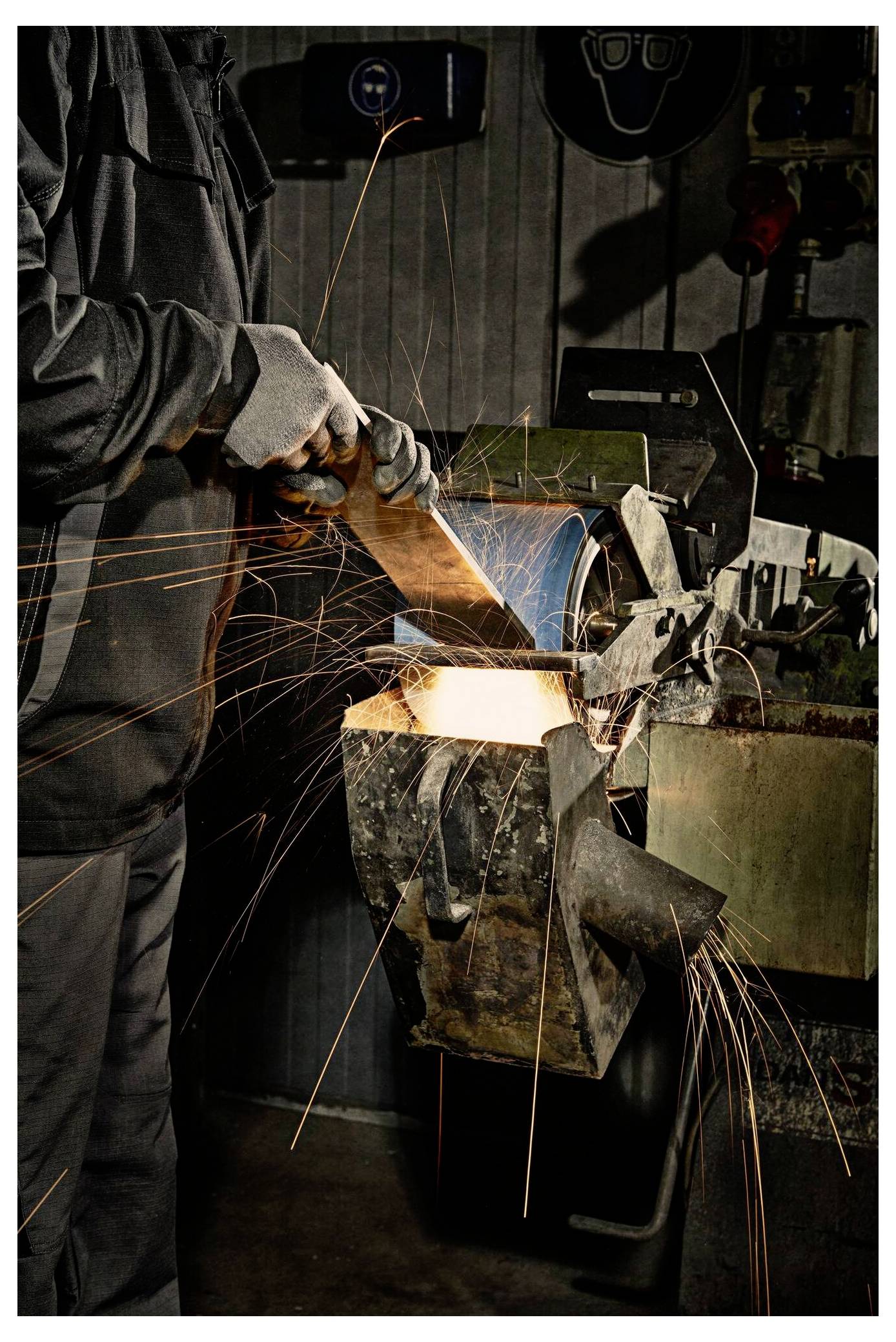 Osoba szlifuje element metalowy na stole szlifierskim, wytwarzając snop iskier. Ma na rękach rękawice ochronne i znajduje się w pomieszczeniu warsztatowym.
