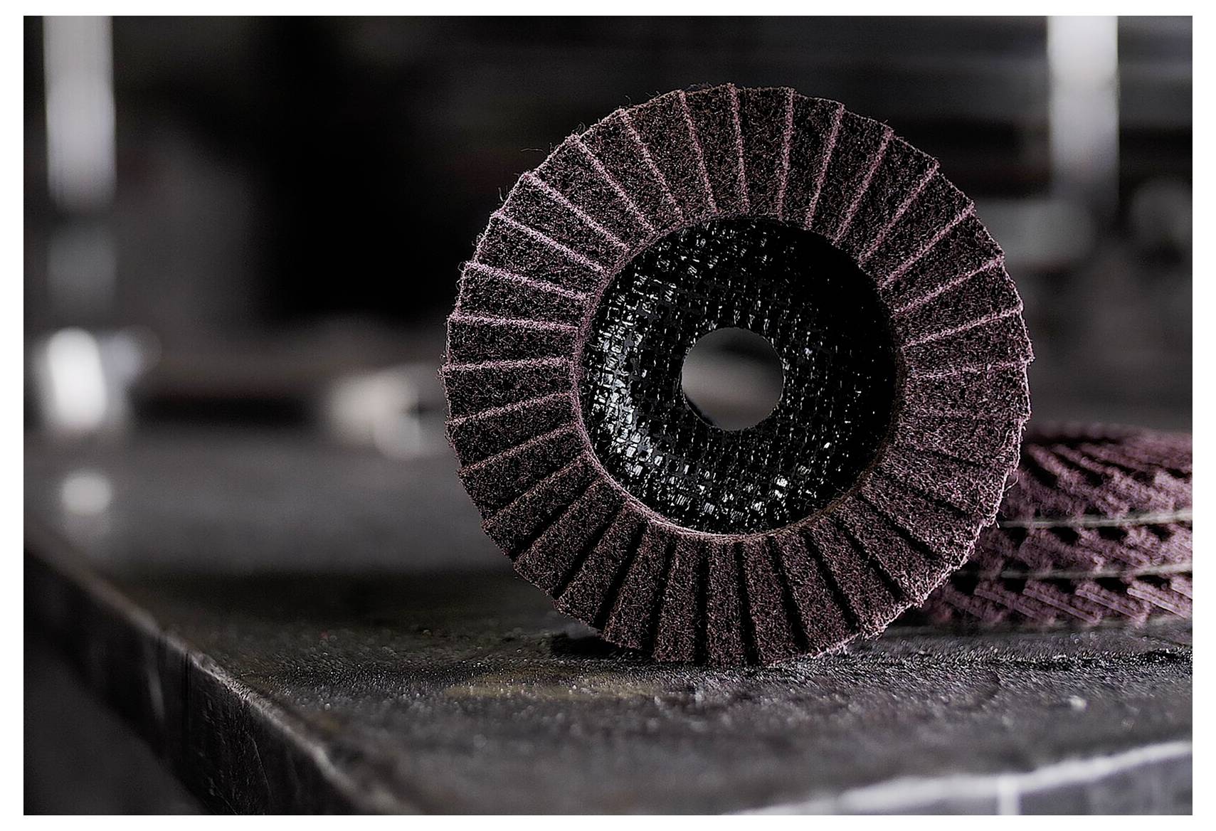 Zbliżenie tarczy szlifierskiej z otworem w centrum, spoczywającej na stole warsztatowym. Widoczna wzorzysta powierzchnia ścierna, używana do wygładzania materiałów.