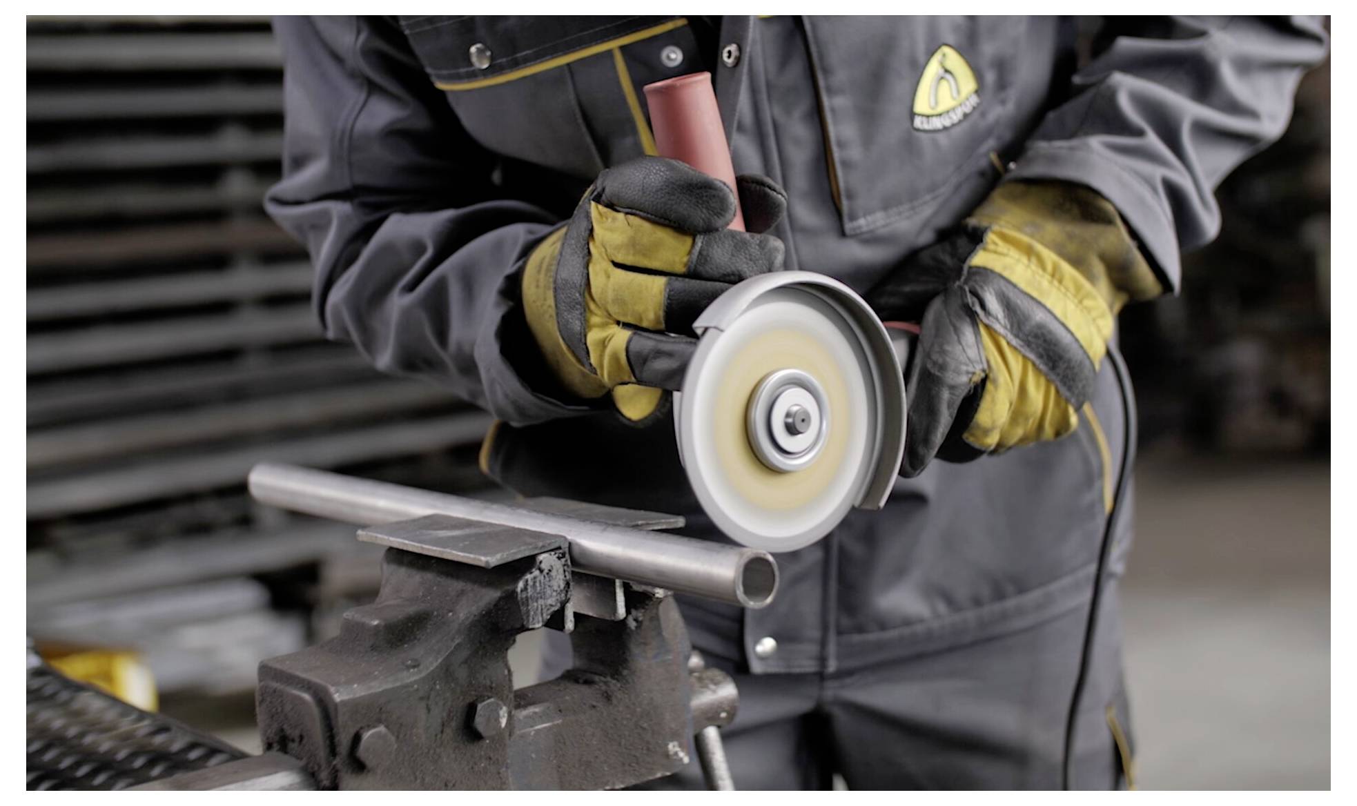 Osoba ubrana w sprzęt ochronny używa szlifierki kątowej do cięcia metalowej rury zamocowanej w imadle, demonstrując proces obróbki metalu.