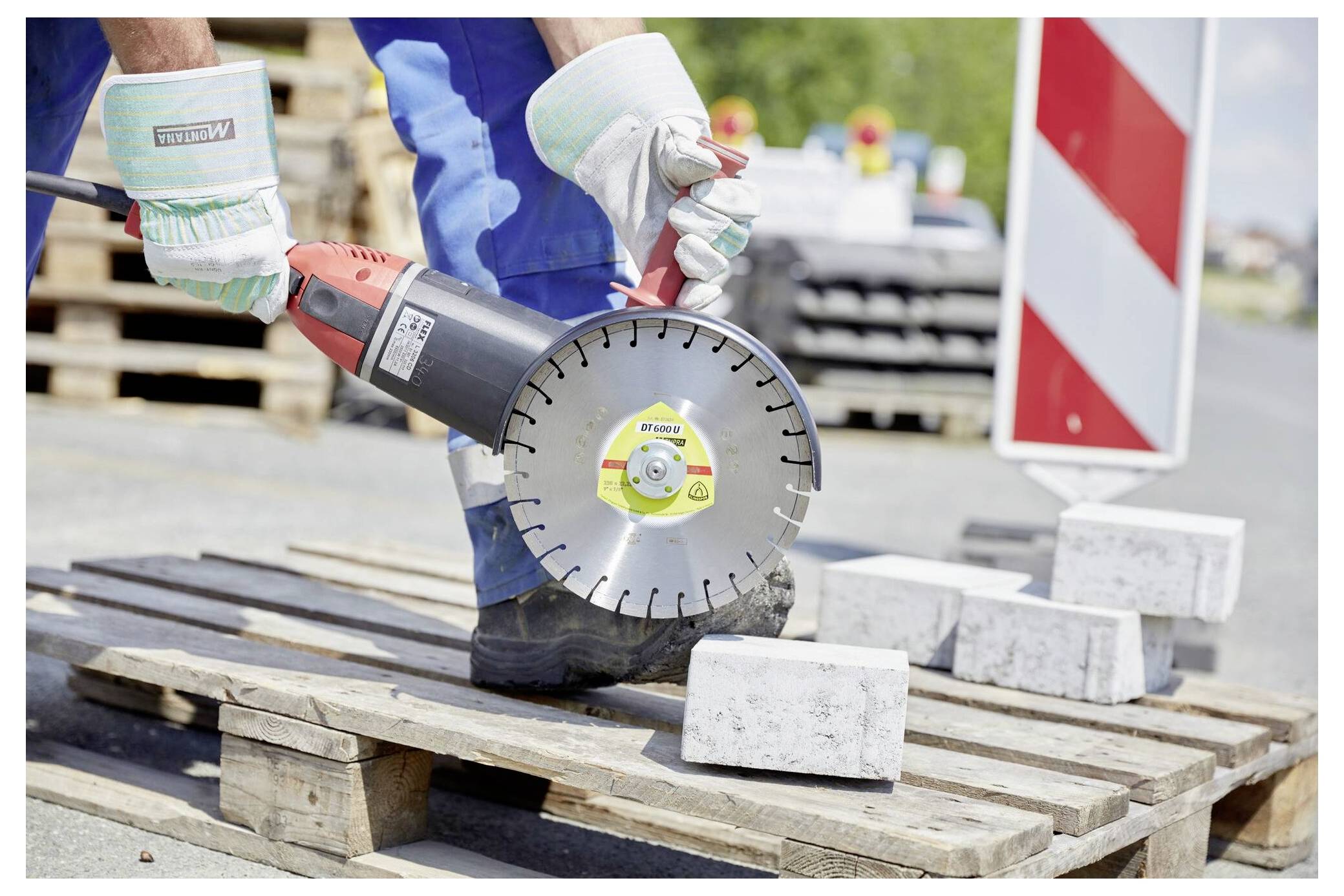 Pracownik w rękawicach i niebieskich kombinezonach używa piły mechanicznej do cięcia bloku betonowego na drewnianej palecie na terenie budowy, a w tle widoczne są znaki bezpieczeństwa.