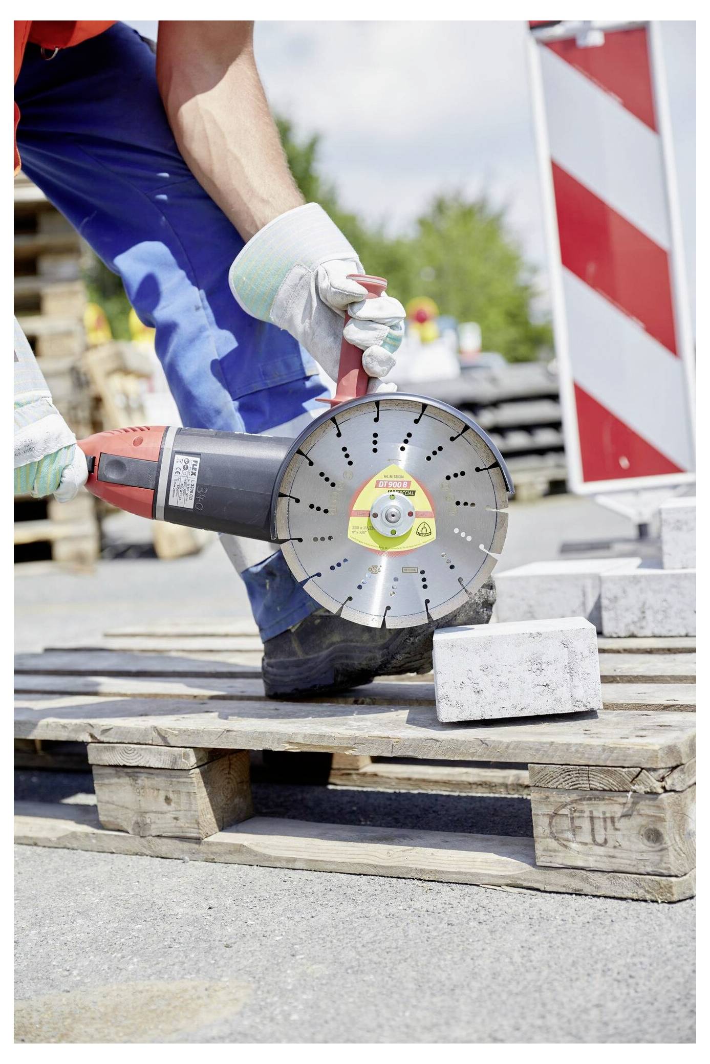 Robotnik budowlany w kamizelce ochronnej i kasku przecina blok betonowy elektryczną piłą na drewnianej palecie na terenie budowy.