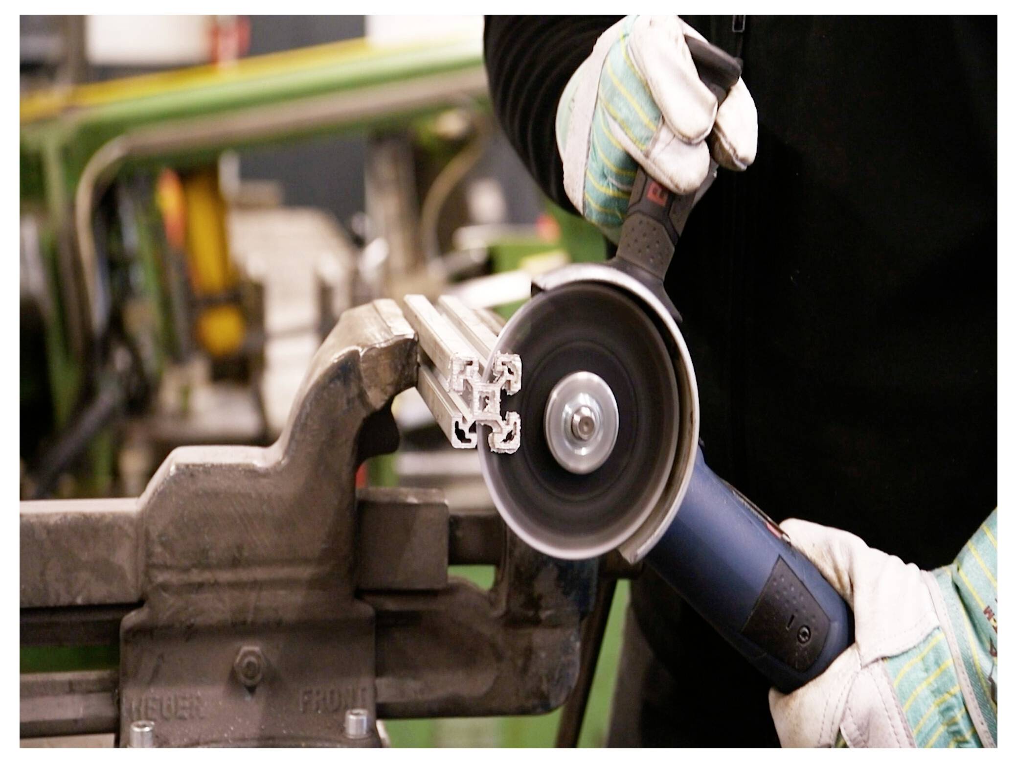 Osoba w rękawicach używa elektronarzędzia do cięcia kawałka metalu, który jest pewnie zamocowany w imadle w przemysłowym otoczeniu.