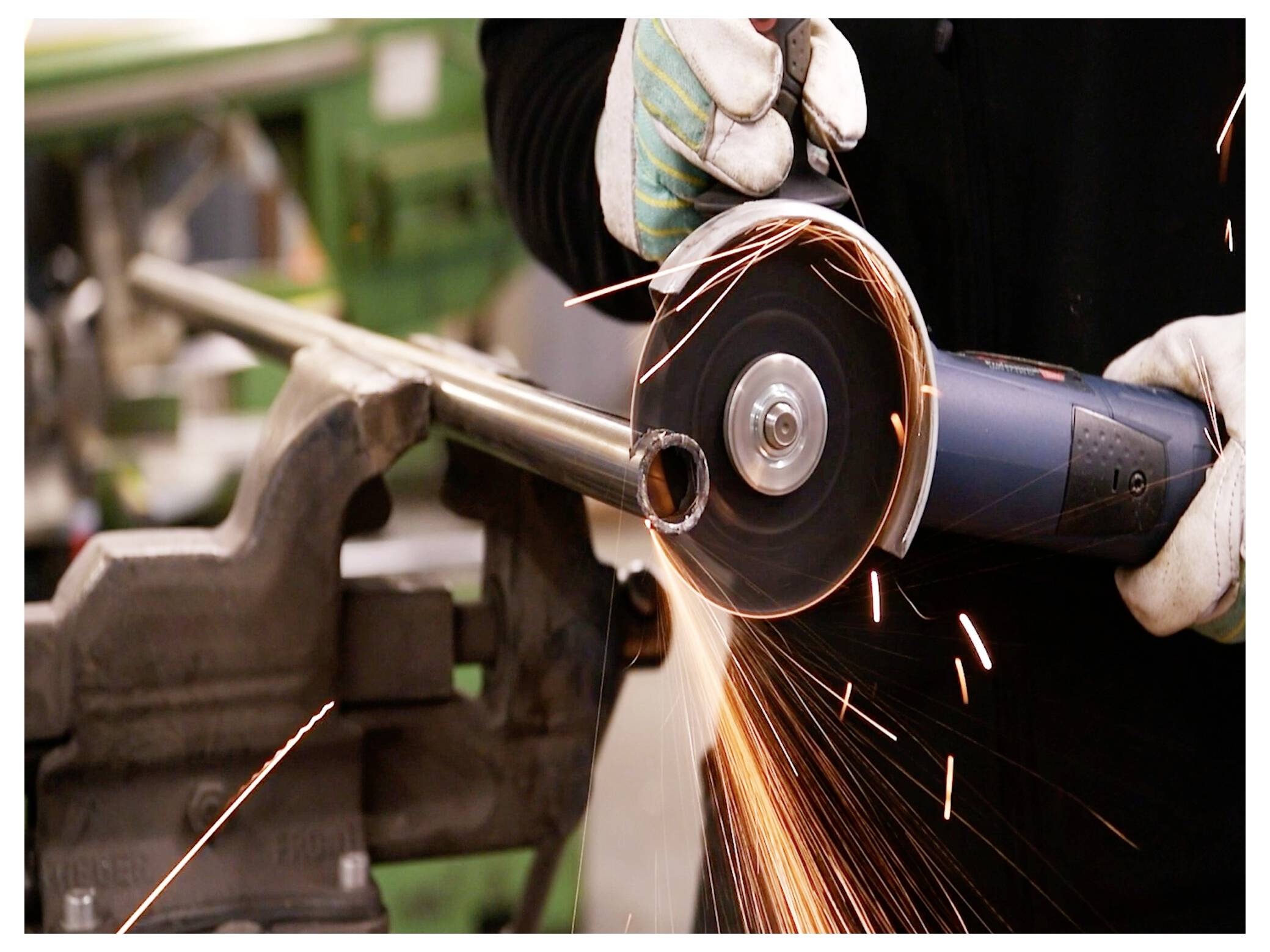 Osoba używa szlifierki kątowej do cięcia metalowej rury, z lecącymi iskrami. Rura jest przytrzymywana przez imadło na stole warsztatowym.