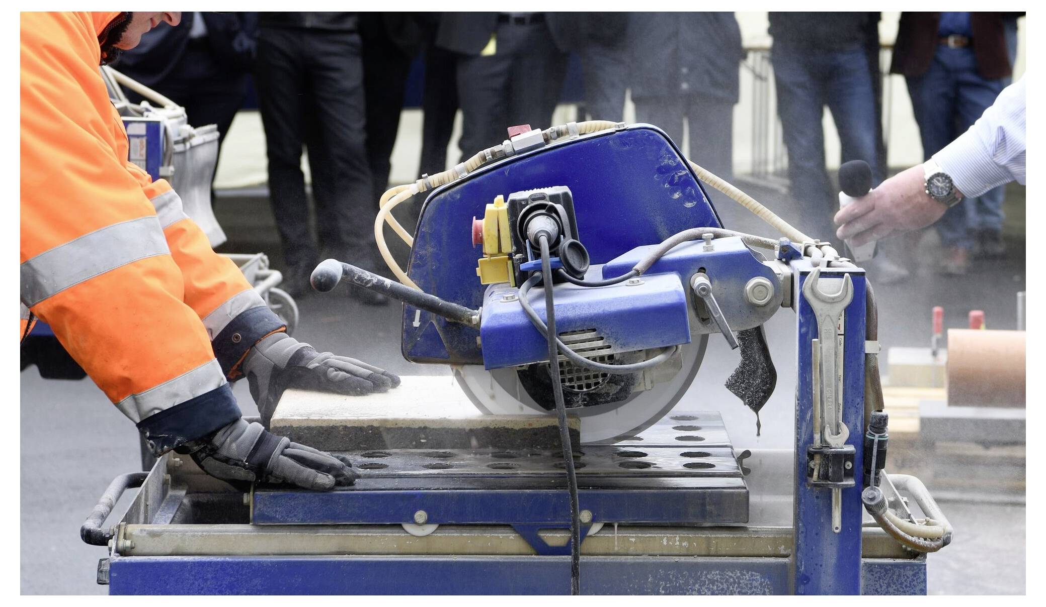 Osoba ubrana w rękawice ochronne i pomarańczową kurtkę obsługuje maszynę do cięcia kamienia, podczas gdy inna osoba trzyma w pobliżu mikrofon.