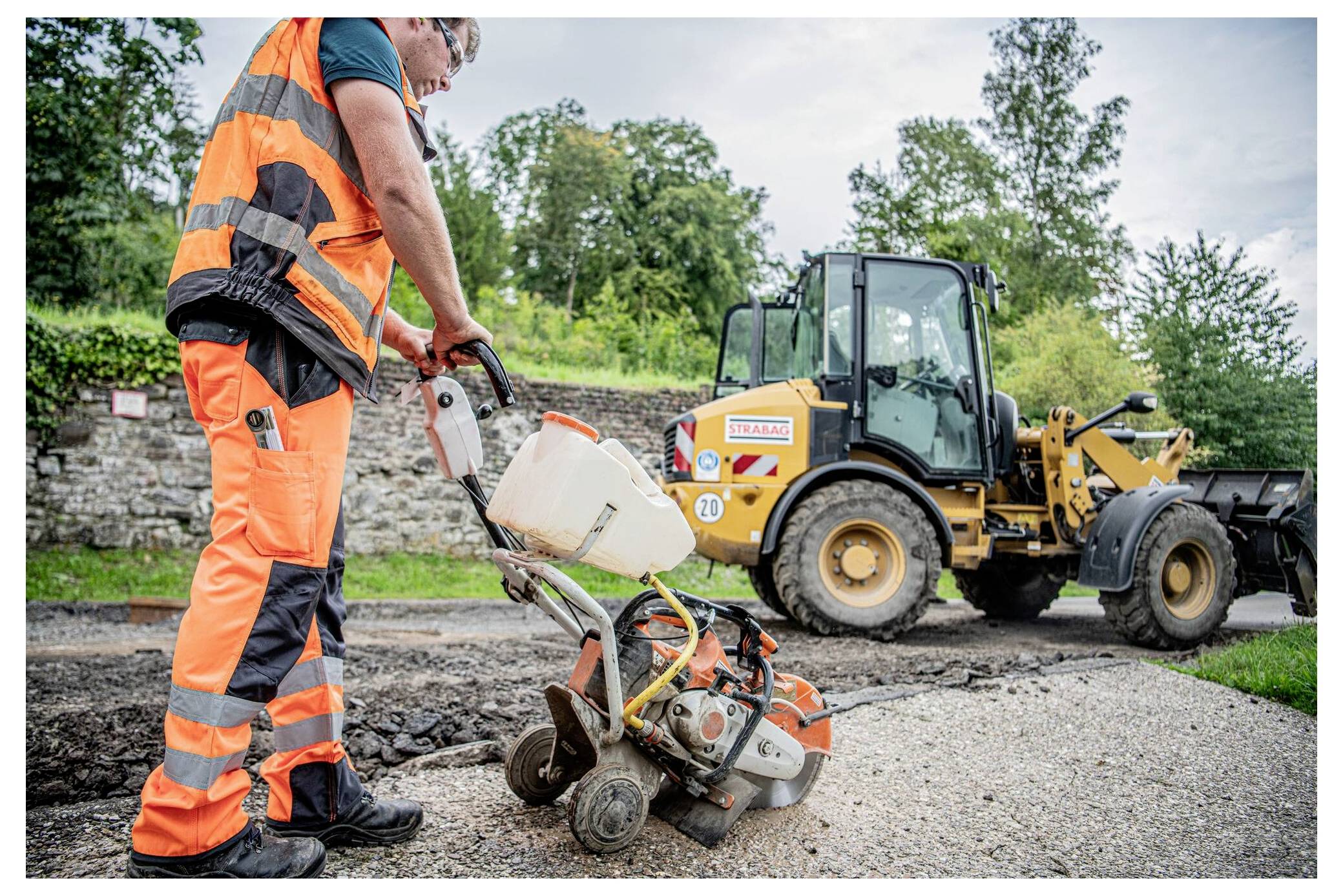 Robotnik budowlany obsługuje maszynę na nawierzchni drogi, z koparką w tle, wśród drzew i kamiennego muru.