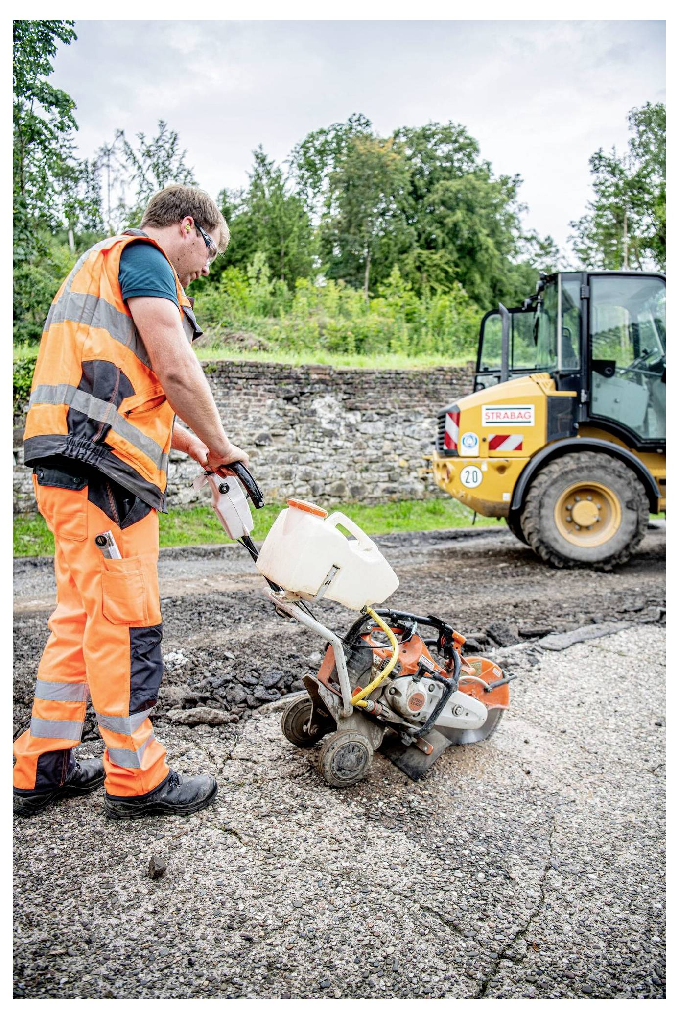 Robotnik budowlany w pomarańczowym stroju ochronnym używa piły do cięcia nawierzchni na uszkodzonym chodniku, podczas gdy ładowarka jest zaparkowana w pobliżu, otoczona zielenią.