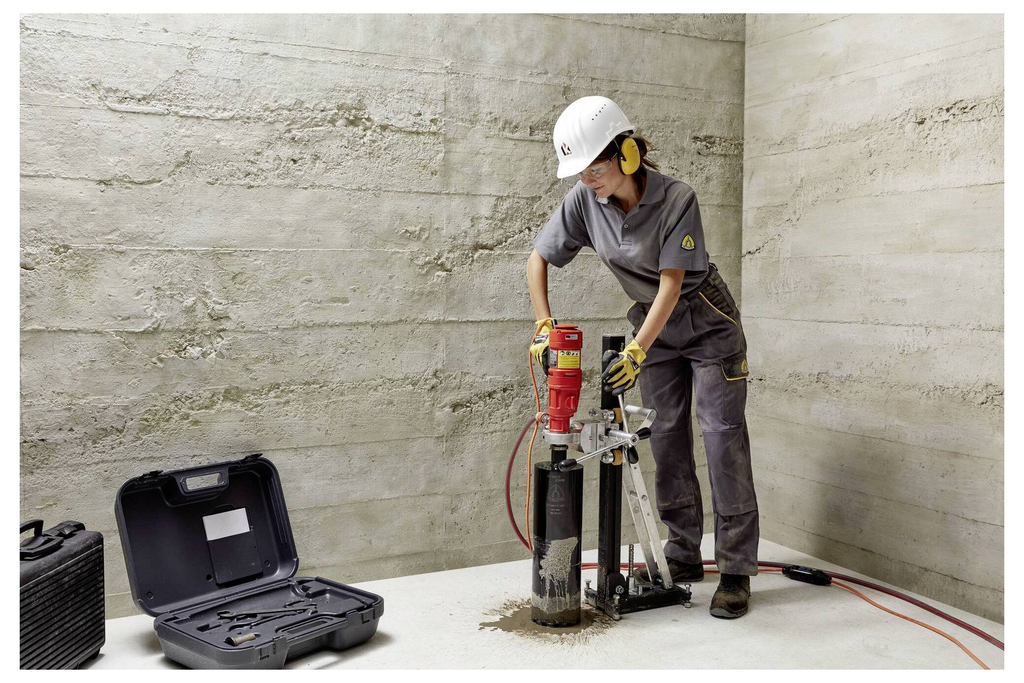 Osoba w sprzęcie ochronnym używa czerwonej wiertarcy rdzeniowej na betonowej podłodze w pustym pomieszczeniu. Czarna walizka na sprzęt jest otwarta na ziemi w pobliżu.