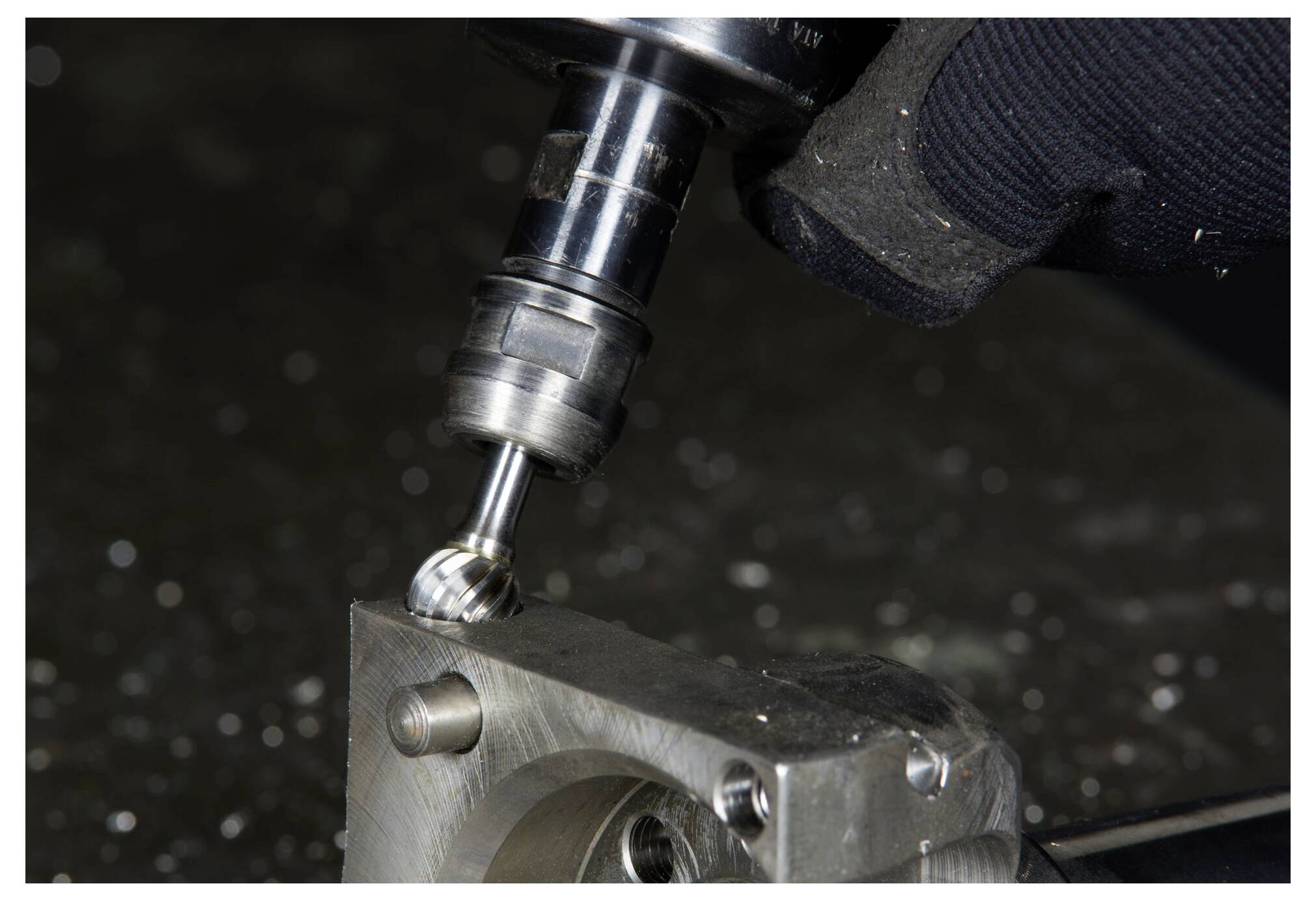 Wiertarka do obróbki metalu tworzy szczegółowy wzór na bloku metalowym przytrzymywanym stabilnie przez dłoń w rękawicy, z widocznymi wiórkami metalu.
