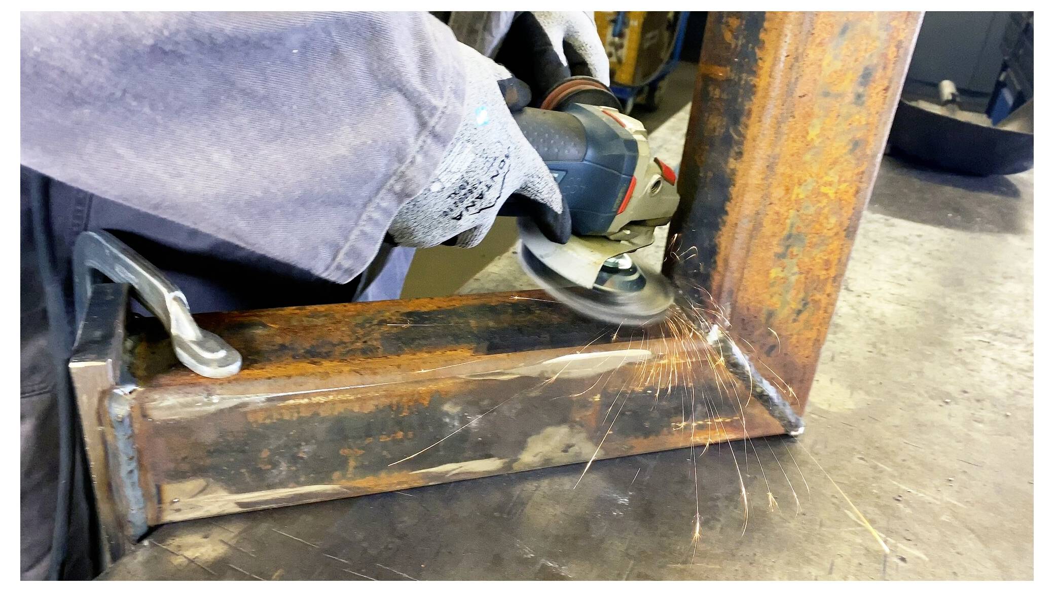 Osoba w rękawicach używa szlifierki kątowej na metalowej ramie, powodując powstawanie iskier. Praca wydaje się odbywać w warsztacie.