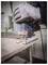 Osoba w rękawicach używa wyrzynarki do cięcia drewnianej deski. Widoczny jest pył drzewny, a obraz oddaje precyzyjne wykonywanie cięcia.