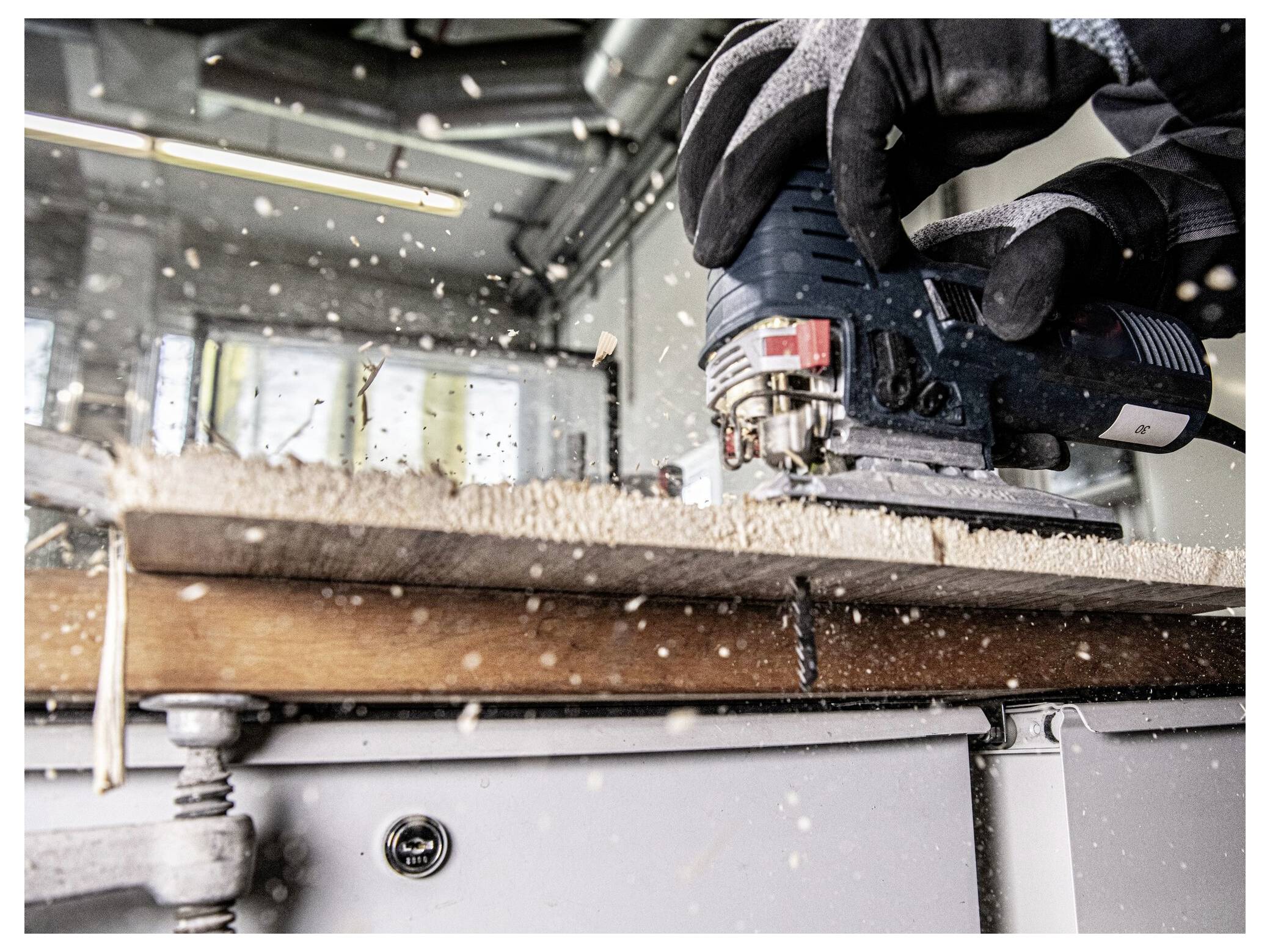 Osoba używająca piły elektrycznej do cięcia drewnianej deski, ubrana w rękawice. Wióry drewniane są widoczne w powietrzu, co wskazuje na wykonywaną czynność.