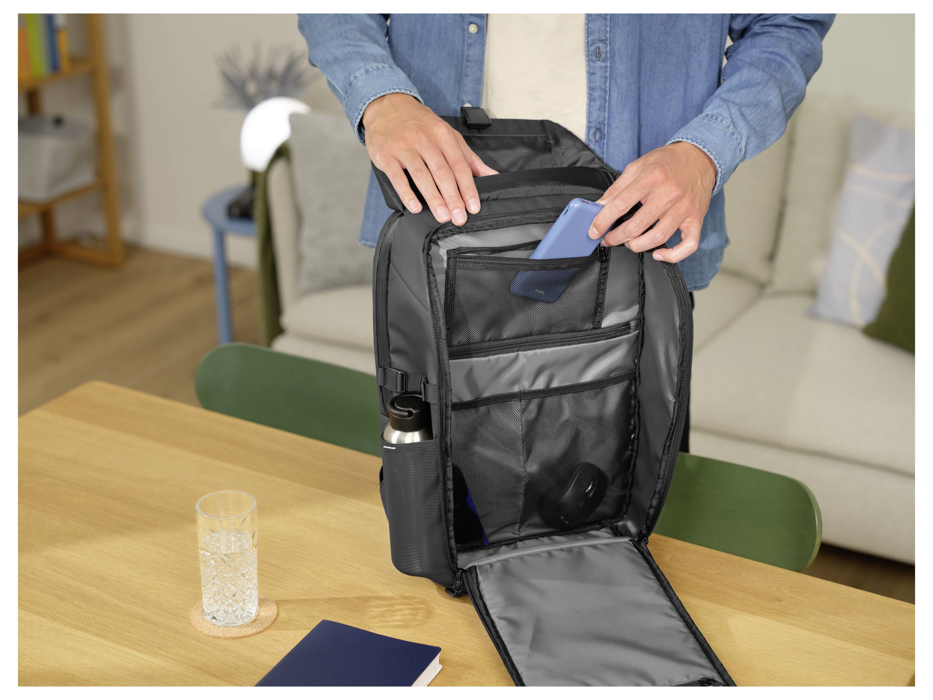 Osoba stoi przy stole, otwierając czarny plecak, aby włożyć do niego niebieski przedmiot. Butelka z wodą znajduje się w bocznej kieszeni, a na stole leży szklanka i notes.