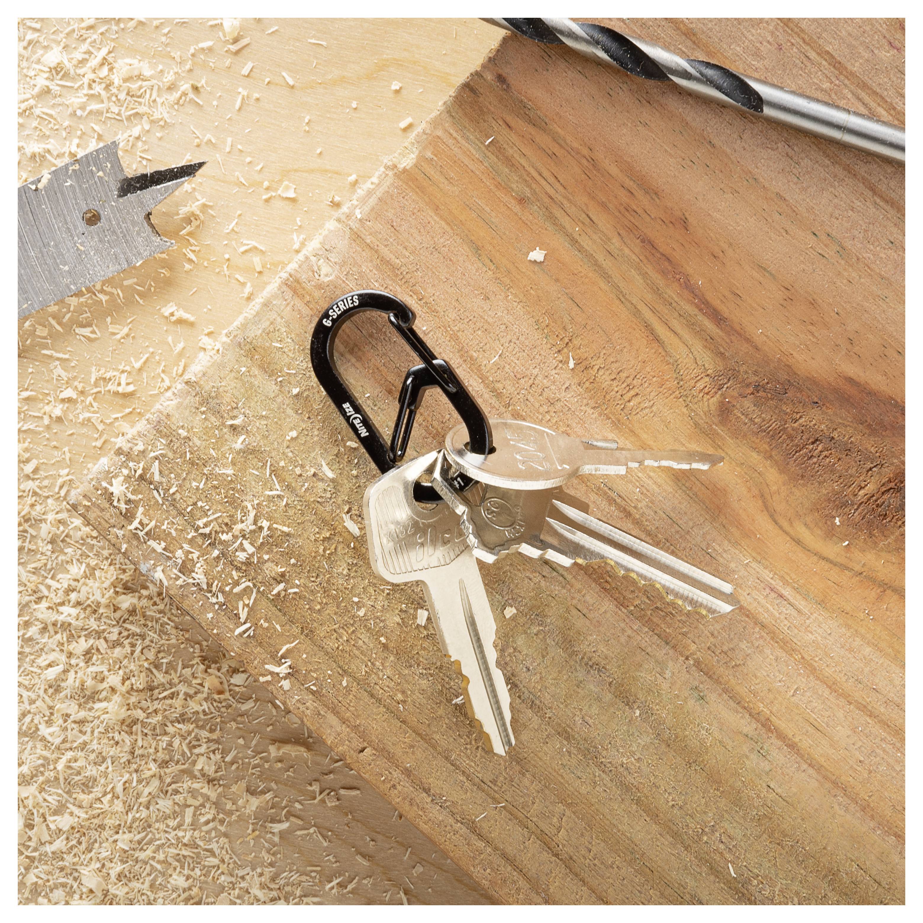 Grupa metalowych kluczy przymocowanych do małego czarnego karabińczyka leży na drewnianej powierzchni, otoczona wiórami drewnianymi i wiertłami.