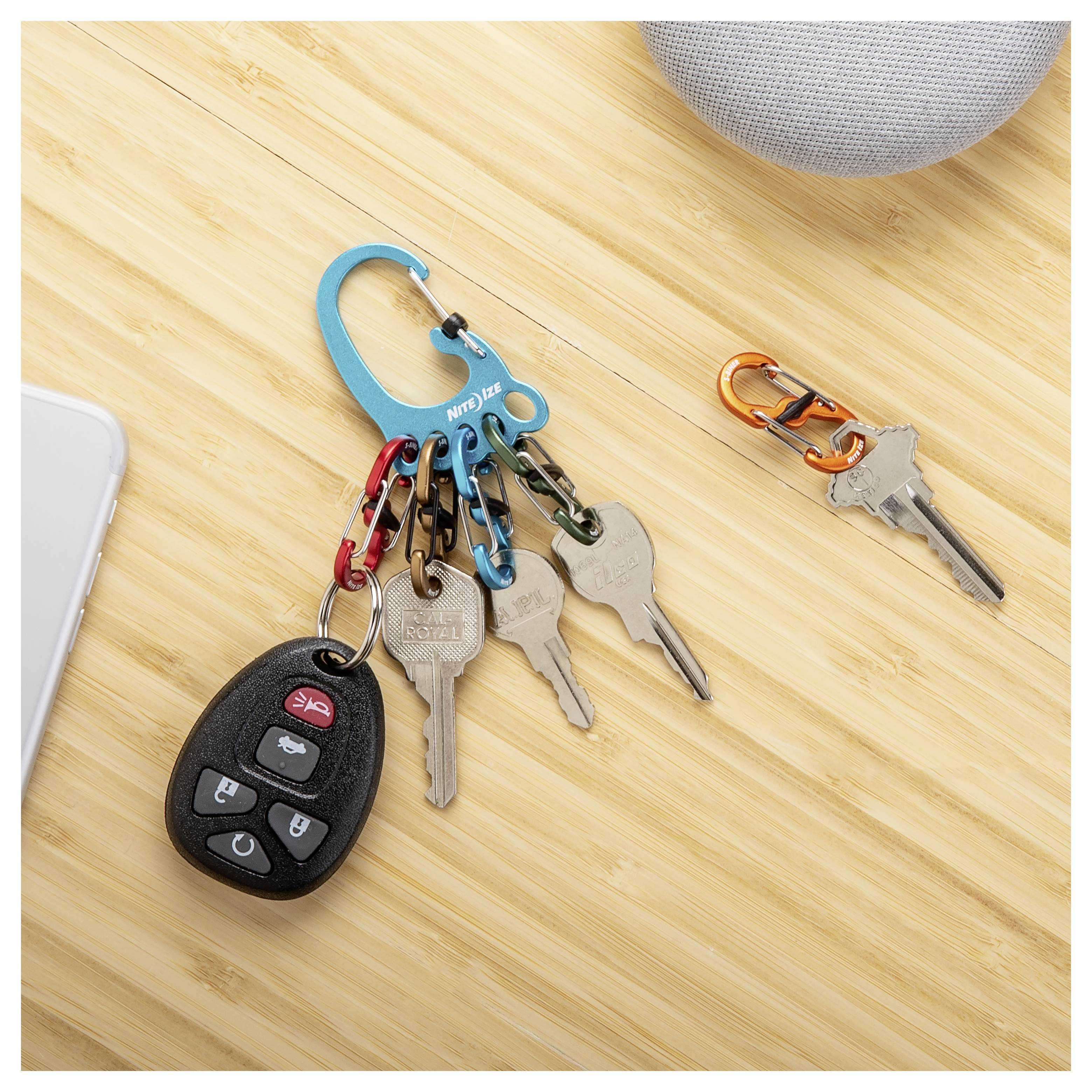Zestaw kluczy, w tym pilot do samochodu, przypięty do niebieskiego karabińczyka na bambusowej powierzchni, obok którego znajduje się mniejszy zestaw kluczy na pomarańczowym karabińczyku.