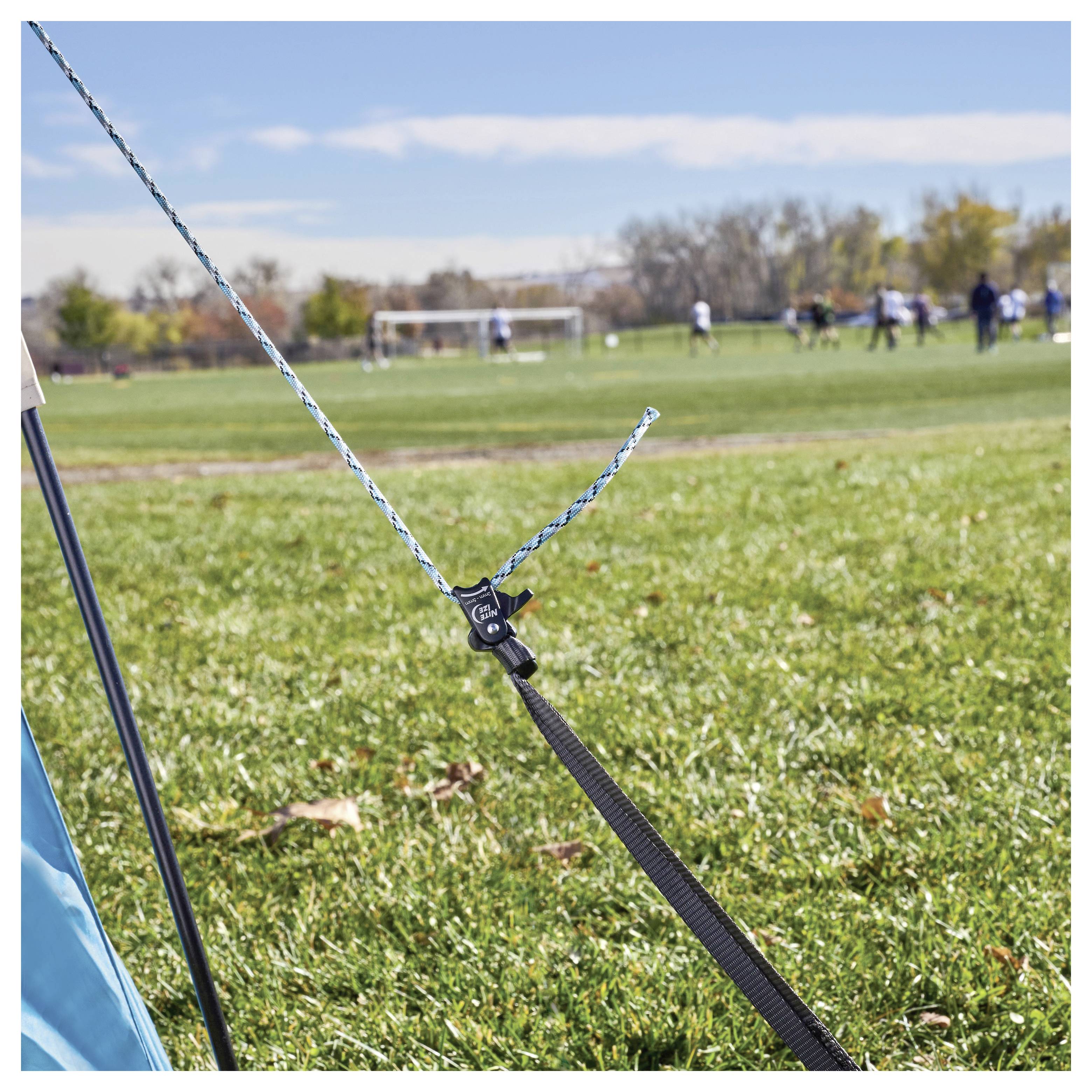 Niebieski namiot rozłożony na trawiastym polu, zabezpieczony liną napinaną za pomocą napinacza. Na tle widoczne są bramki piłkarskie i gracze.