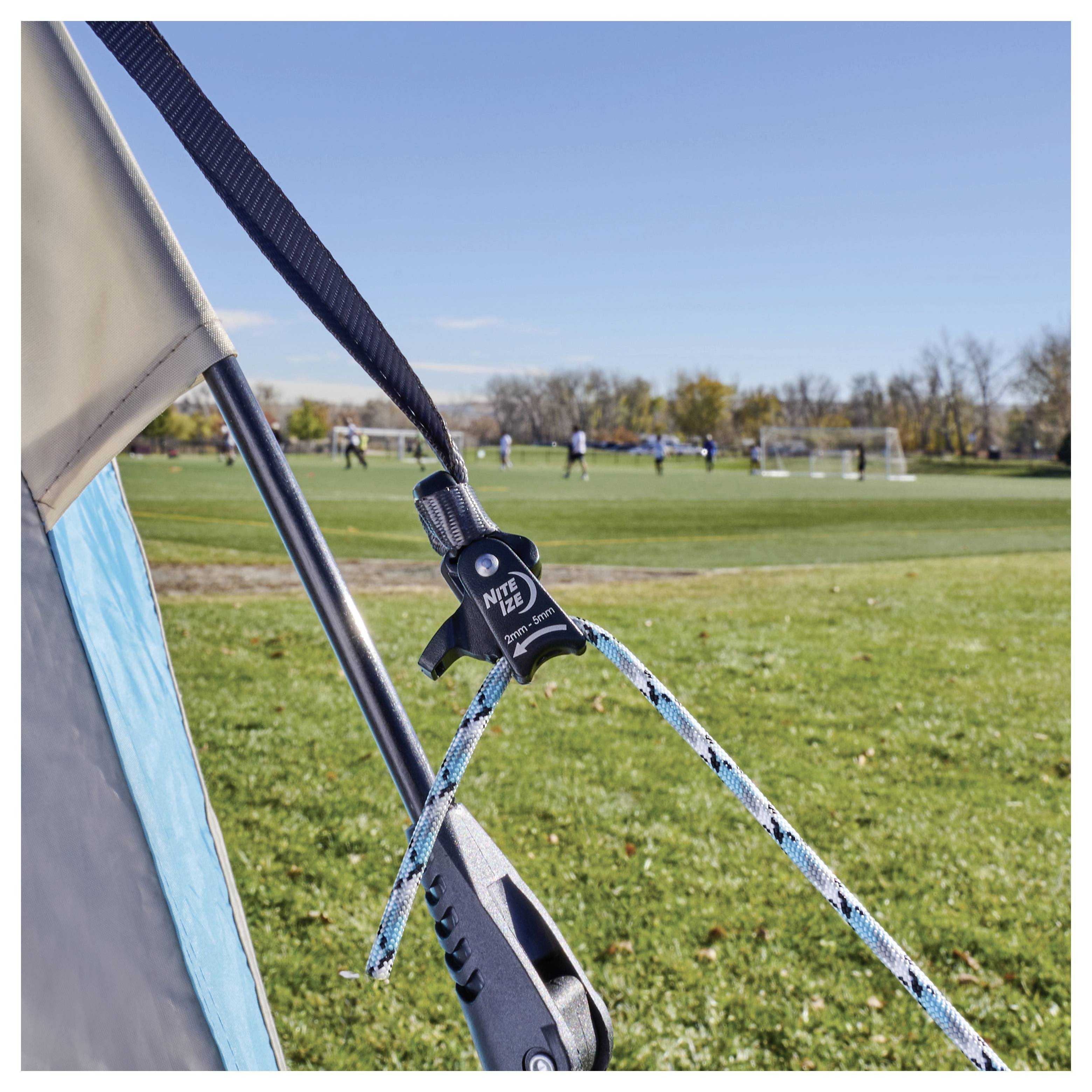 Zbliżenie gwoździa namiotu i liny zabezpieczającej namiot na trawiastym polu, z widocznym meczem piłki nożnej w tle.