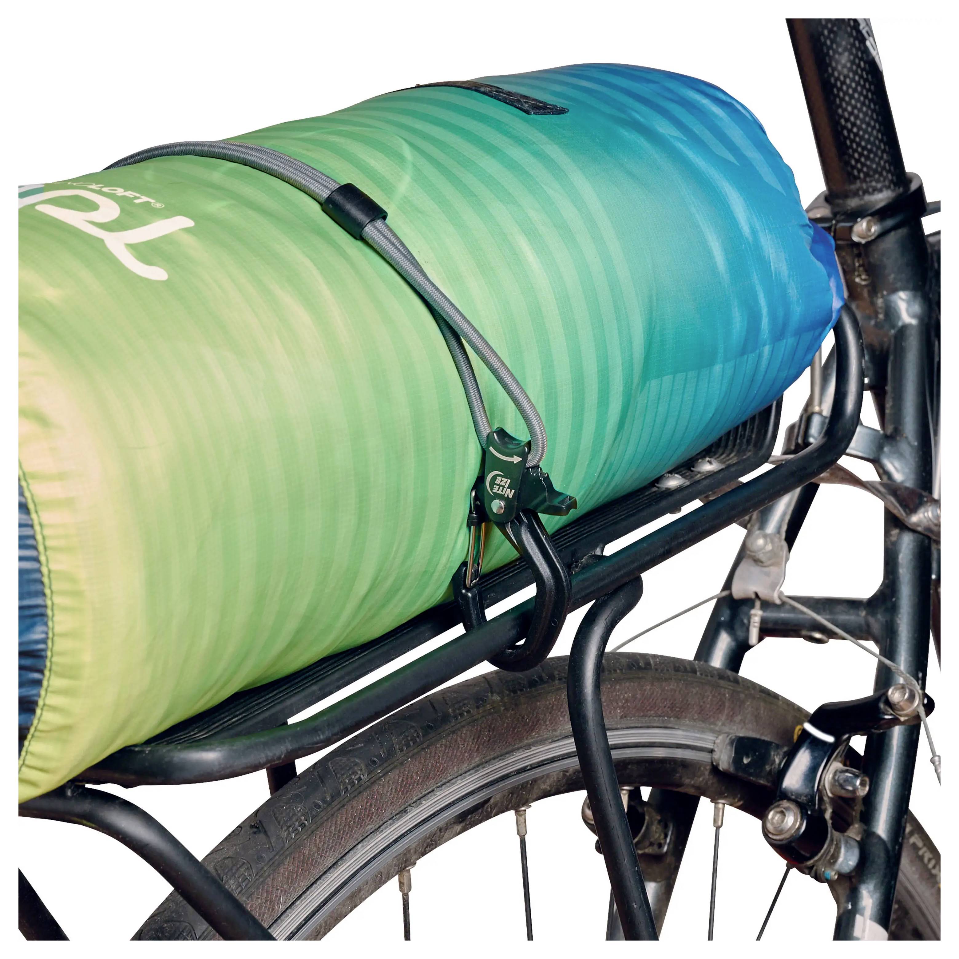 Zwinięty śpiwór w kolorach zielonym i niebieskim jest zamocowany na tylnym bagażniku roweru za pomocą małej kłódki i linki.