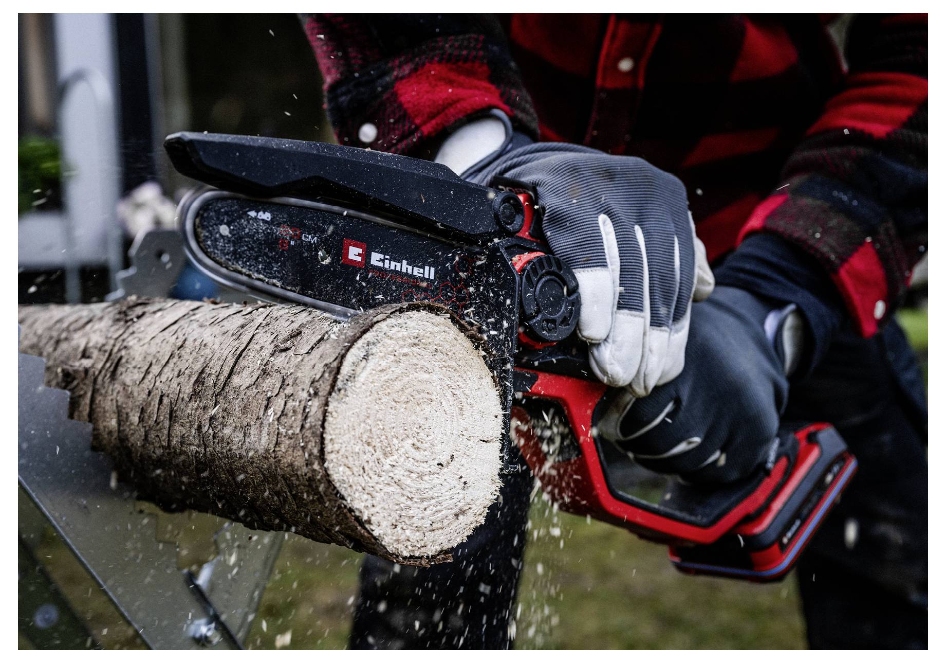 Osoba w ochronnych rękawicach przecina kłodę małą piłą łańcuchową na wolnym powietrzu. Trociny unoszą się w powietrzu, gdy piła łańcuchowa przetyka przez drewno.