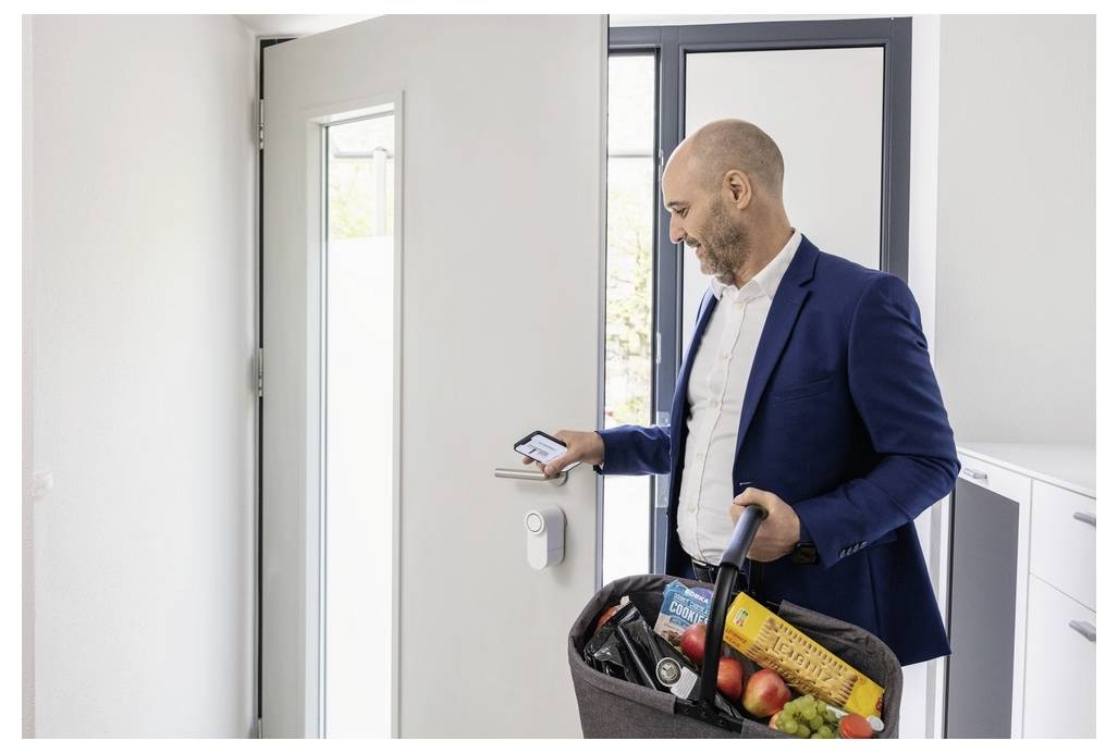 Osoba wchodzi do domu z koszem zakupów, używając smartfona do odblokowania drzwi za pomocą inteligentnego zamka.