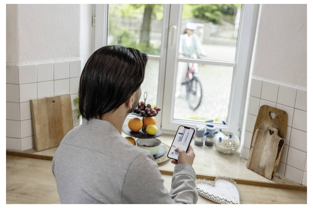 Osoba w kuchni patrzy na smartfona wyświetlającego transmisję wideo. Przez okno widać dziecko jadące na rowerze na zewnątrz.