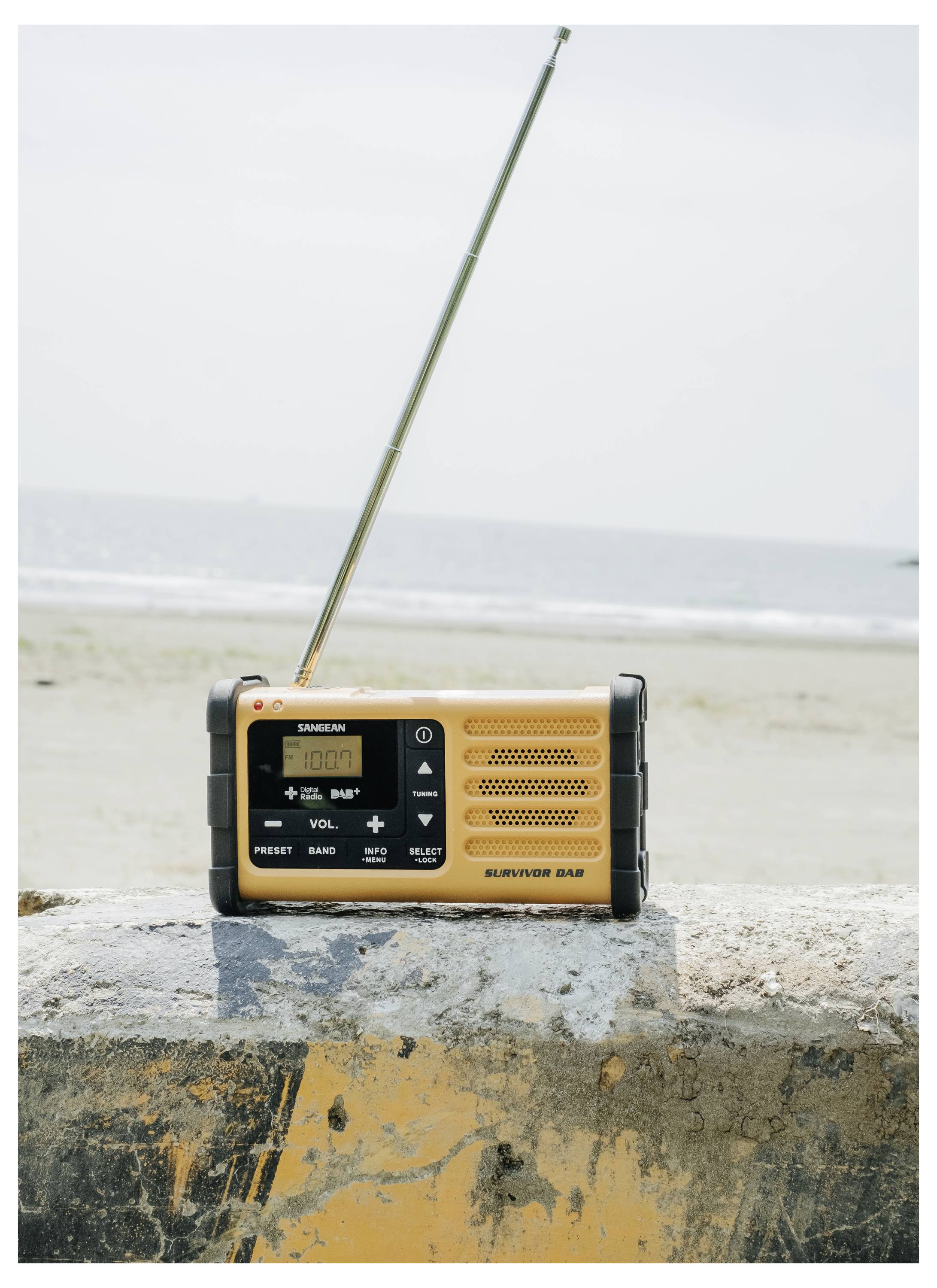 Przenośny, pomarańczowo-czarny radioodbiornik z wysuniętą anteną jest umieszczony na betonowym parapecie nad brzegiem morza, z widokiem na rozległą wodną przestrzeń w tle.