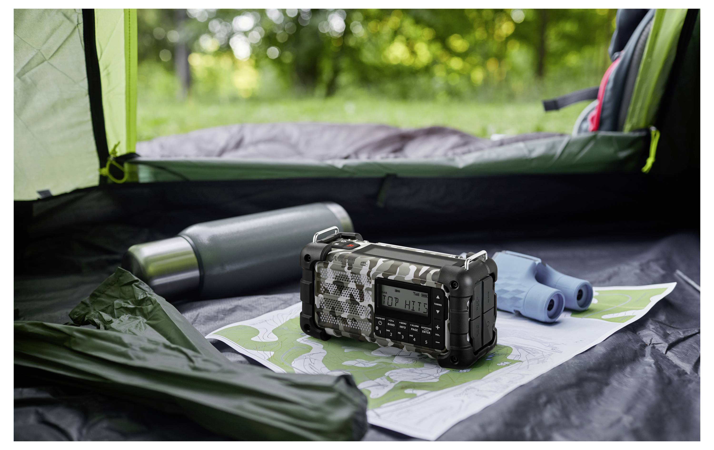 Przenośne radio w kamuflażowym wzorze leży na mapie wewnątrz namiotu, towarzyszą mu termos i lornetka; na zewnątrz widać bujną zieleń.