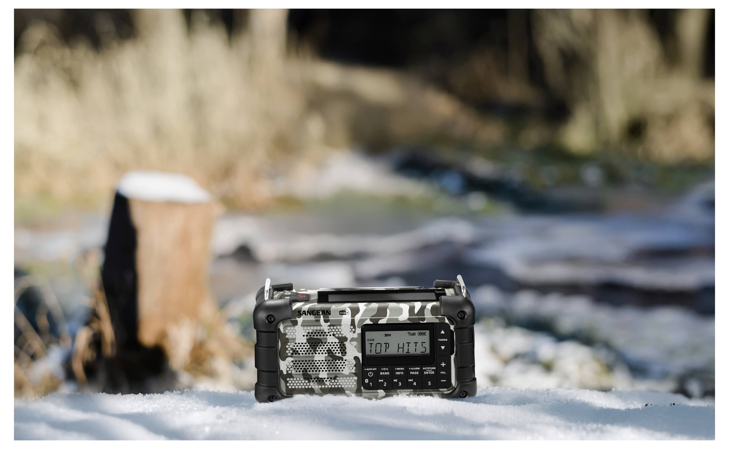 Przenośne radio w kamuflażowym wzorze spoczywa na śniegu, na rozmytym, naturalnym tle z drzewami i pniem.