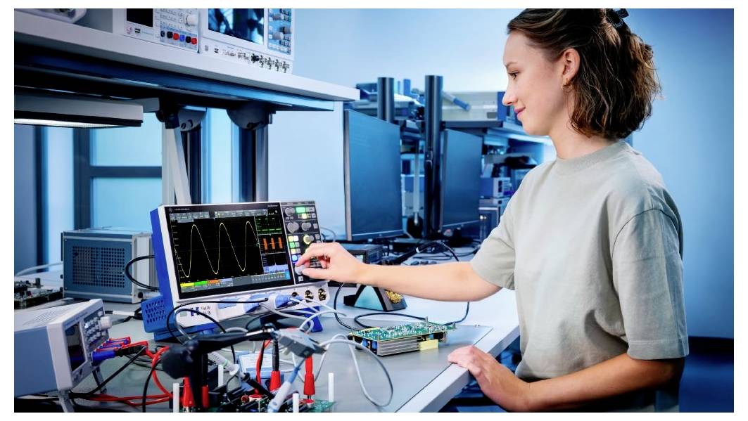 Osoba obsługuje oscyloskop wyświetlający przebiegi fal na stanowisku laboratoryjnym zapełnionym sprzętem elektronicznym, w środowisku zorientowanym na technologię.