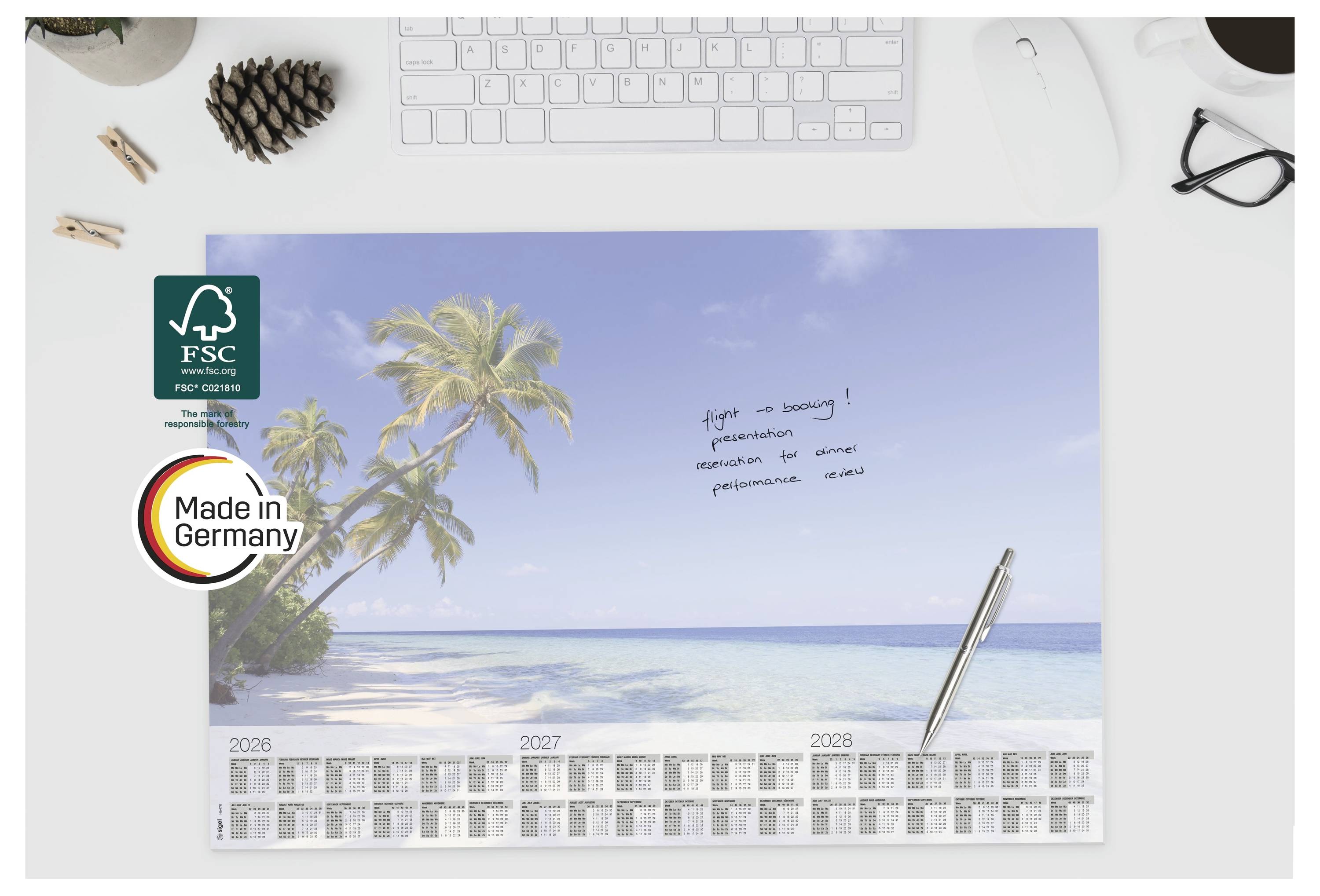 Biurkowy kalendarz przedstawiający tropikalną scenę plażową z palmami i małą notatką napisaną ręcznie; logotypy FSC i "Made in Germany" znajdują się po lewej stronie.