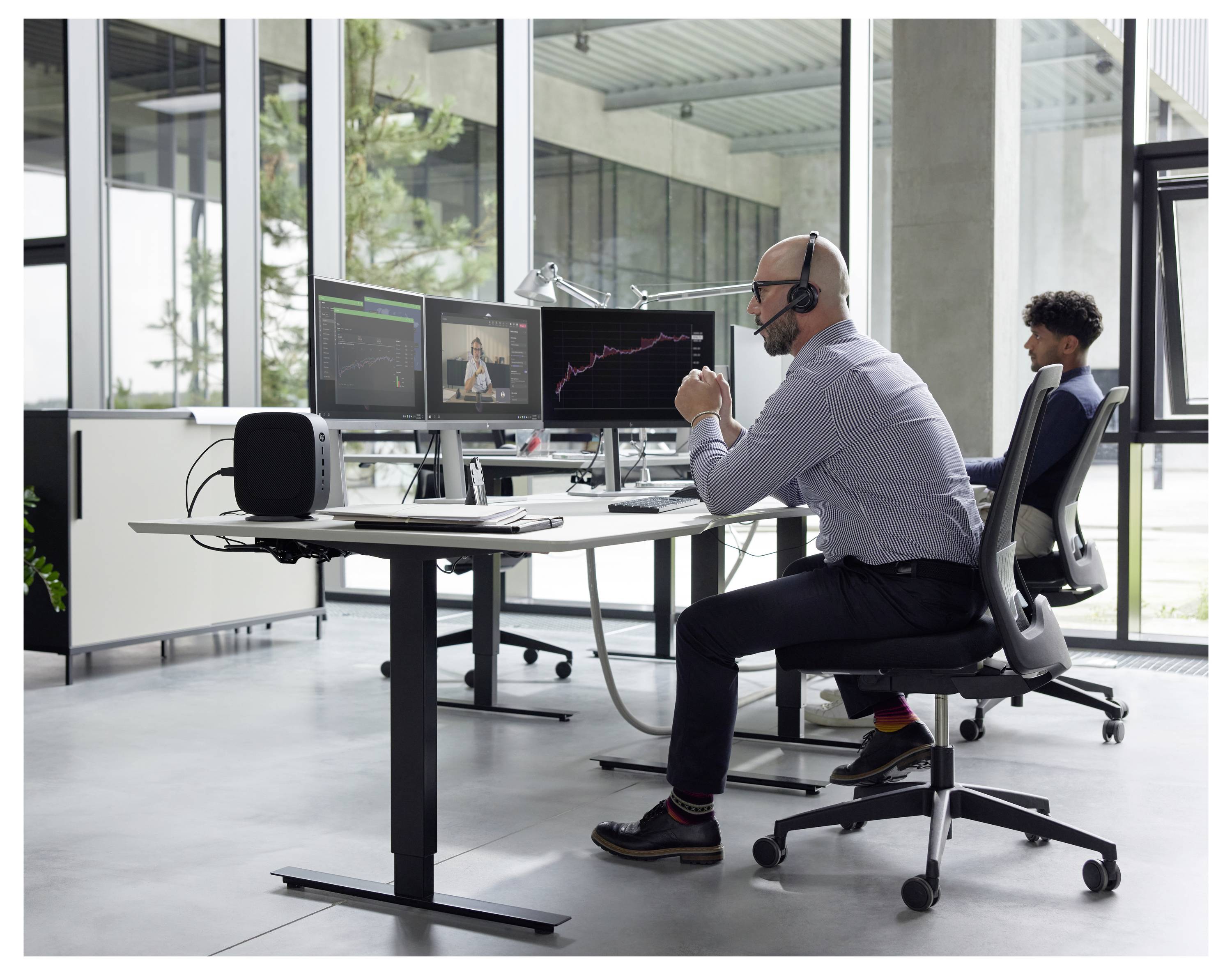 Mężczyzna siedzący przy biurku w słuchawkach uczestniczy w wirtualnym spotkaniu na swoim komputerze, z wieloma ekranami wyświetlającymi wykresy i uczestników.