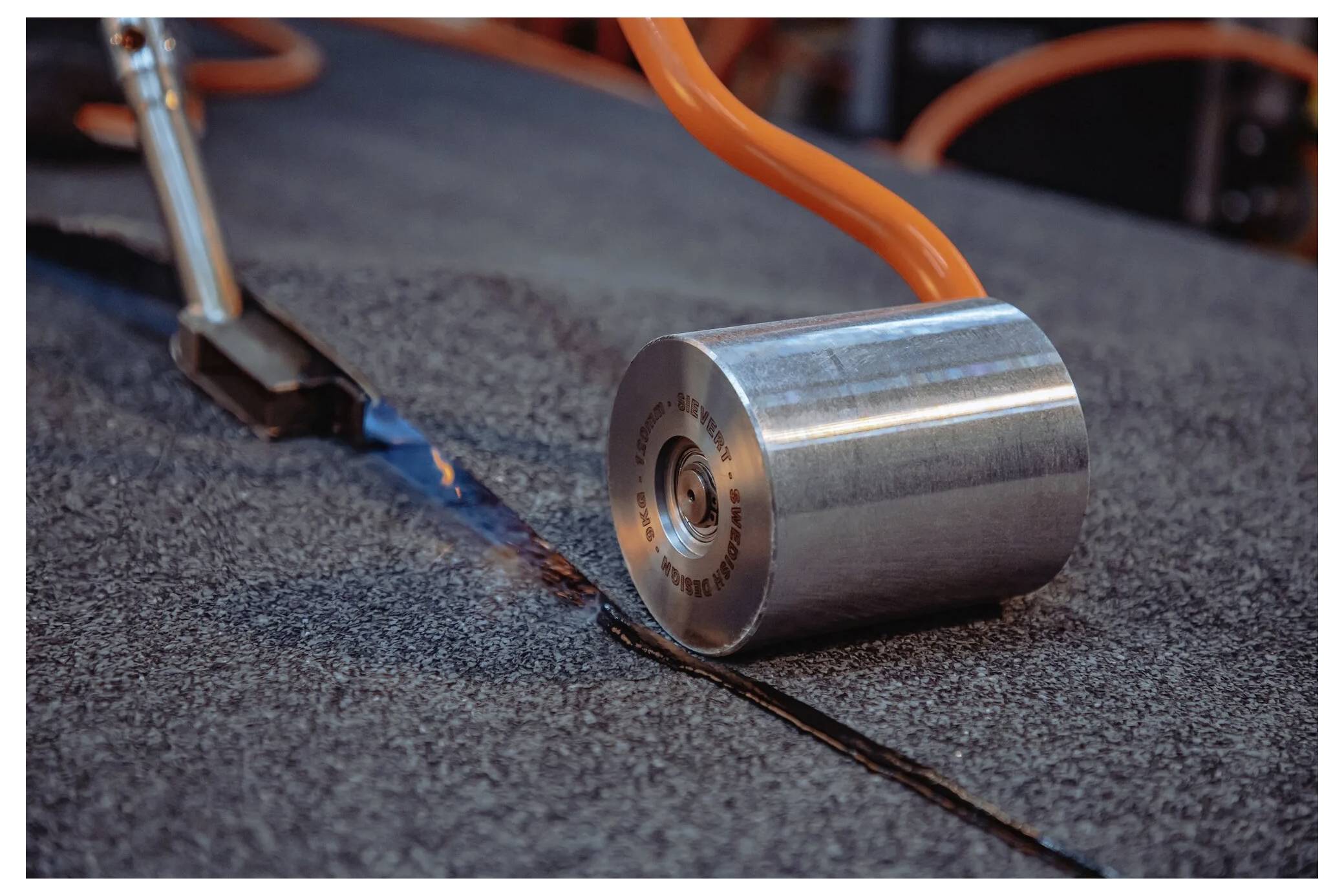 Zbliżenie metalowego wałka naciskającego na szew materiału dachowego, wygładzającego go, z widocznym niebieskim klejem pod spodem.