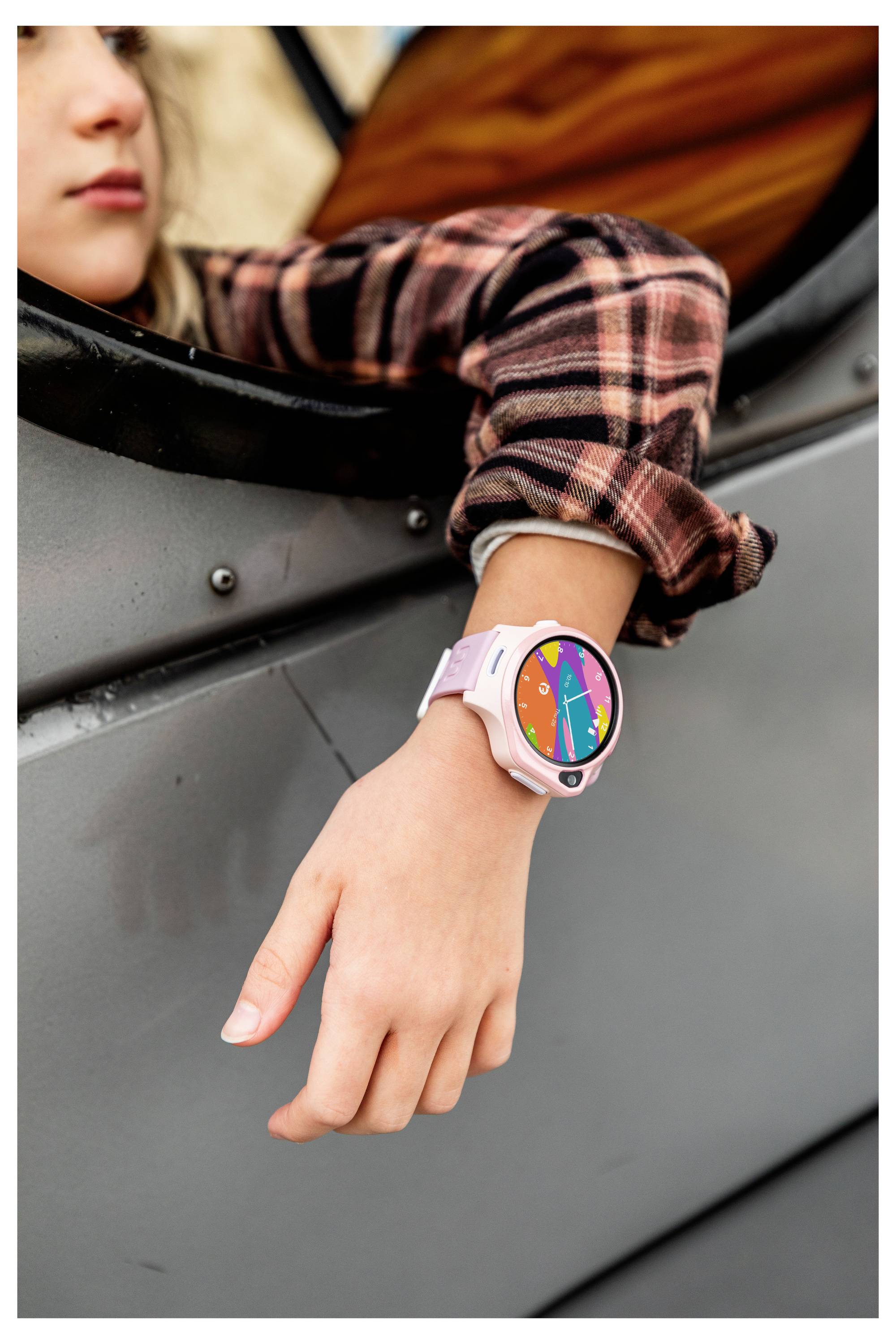 Osoba nosząca różowy smartwatch z kolorowym, geometrycznym wzorem tarczy, opierająca ramię na krawędzi okna pojazdu.