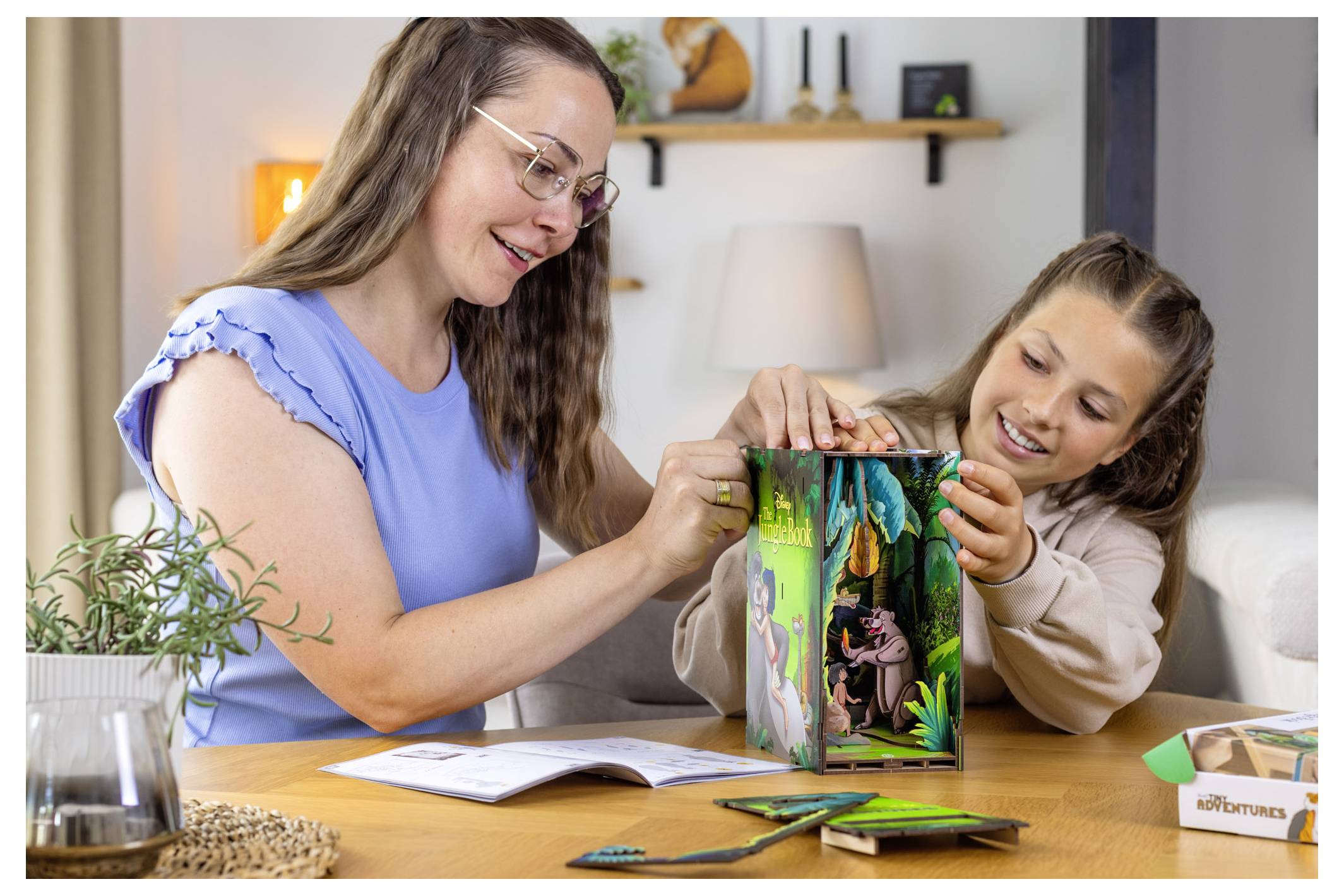Kobieta i dziecko wspólnie składają kolorowy papierowy model sceny dżungli przy stole, oboje uśmiechają się i są całkowicie zaangażowani w zajęcie.