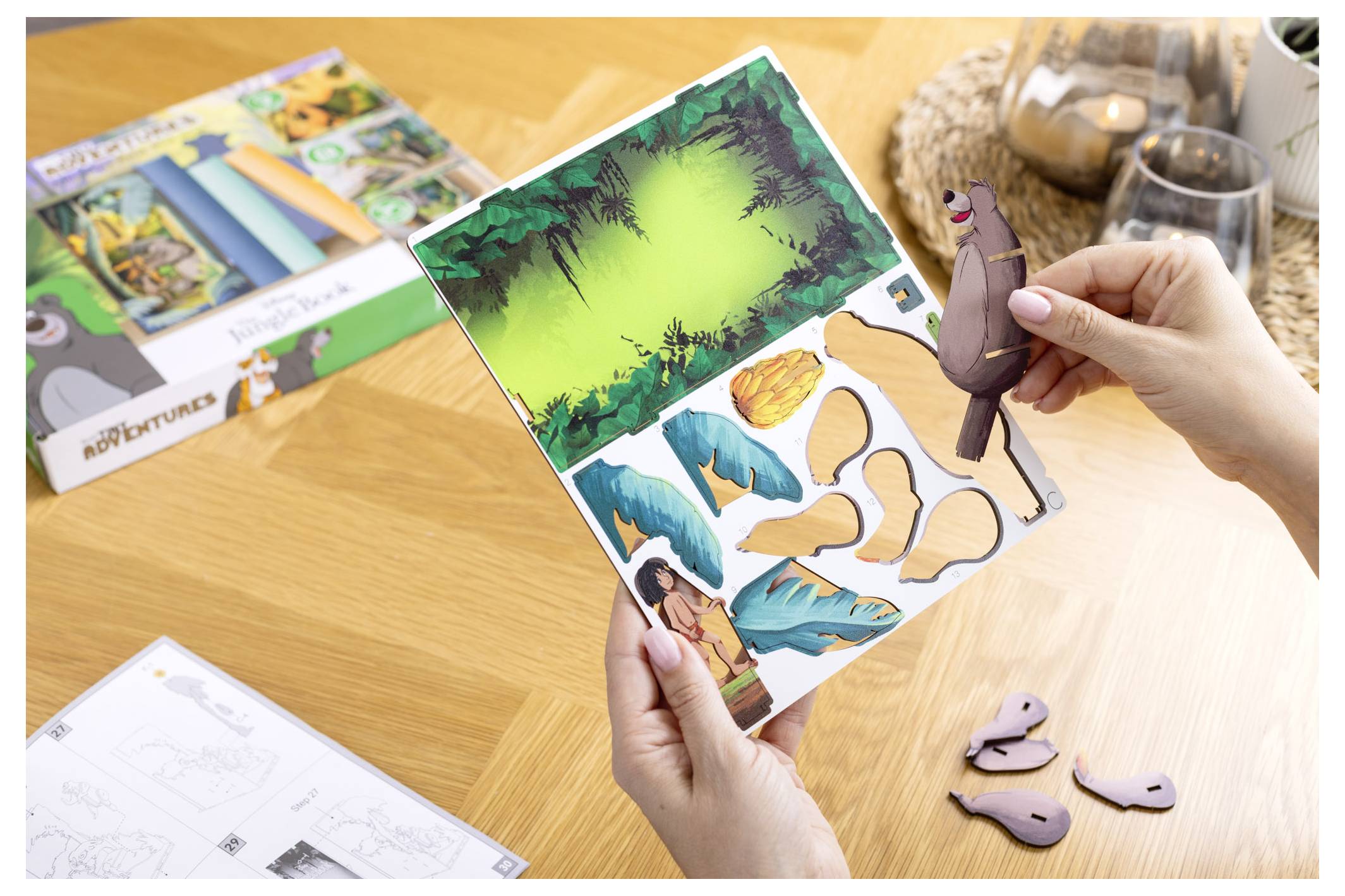 Ręce trzymające arkusz z wyciętymi postaciami z Księgi Dżungli na tle dżungli. Elementy obejmują postać i różne fragmenty kształtów.