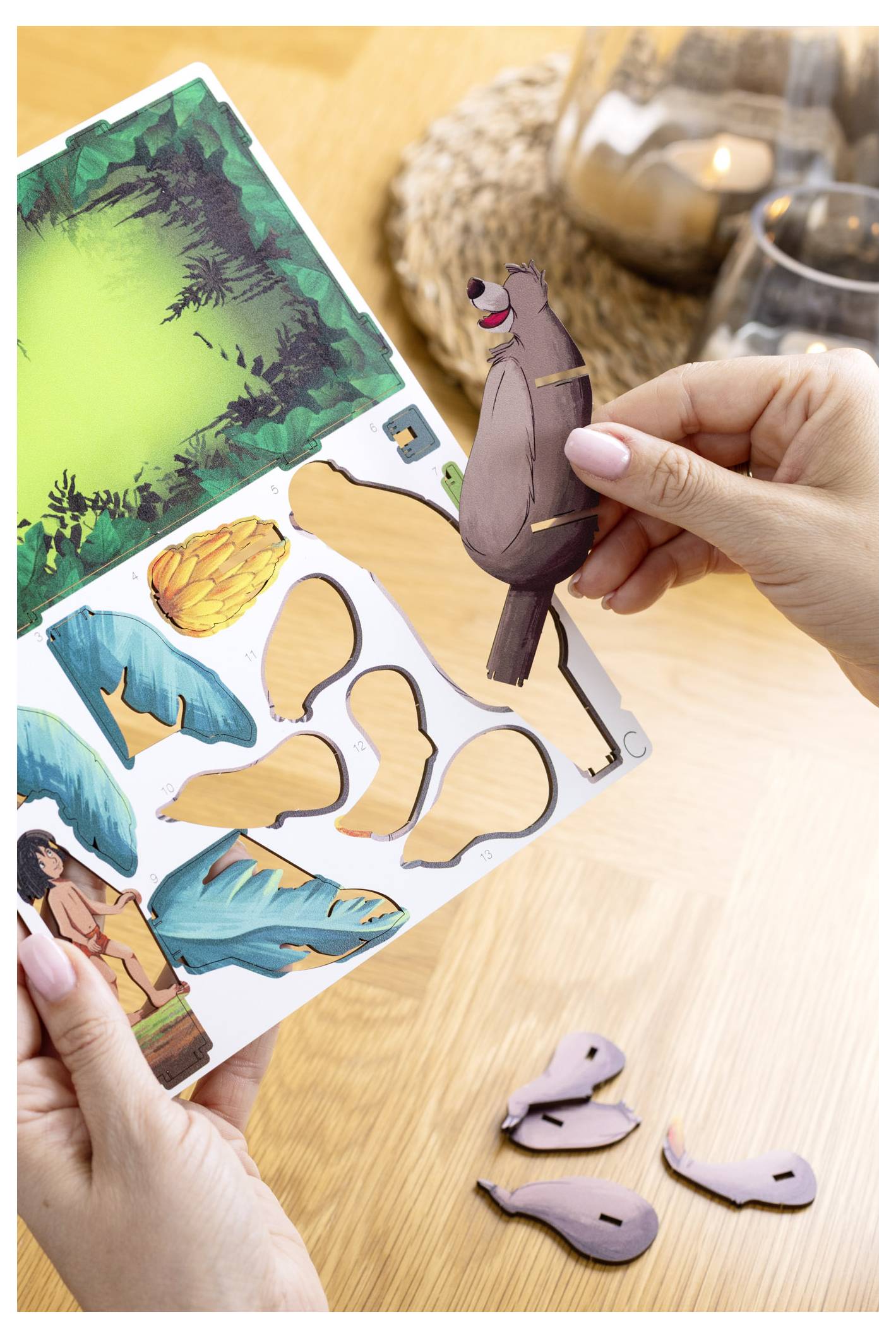 Osoba składa papierową ozdobę przedstawiającą leniwca z kreskówki, używając arkusza do wycinania elementów z motywem dżungli, leżącego na drewnianym stole.