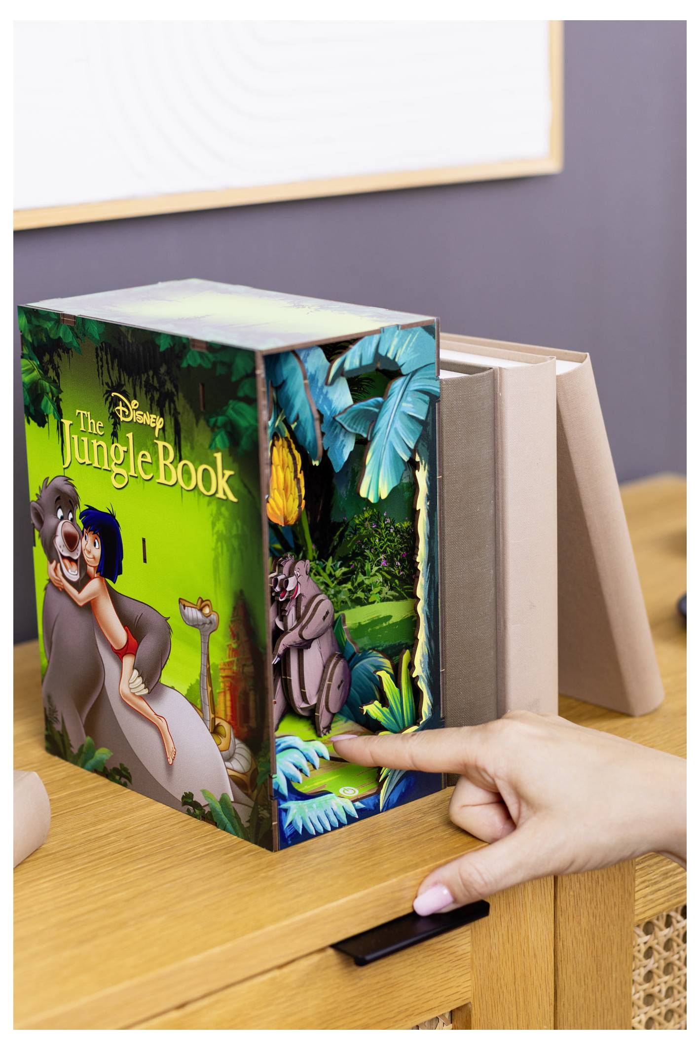 Drewniana półka z kilkoma książkami stoi w salonie. Ktoś wyciąga książkę zatytułowaną 'Księga dżungli', na której okładce widoczne są scenerie dżungli i postacie.