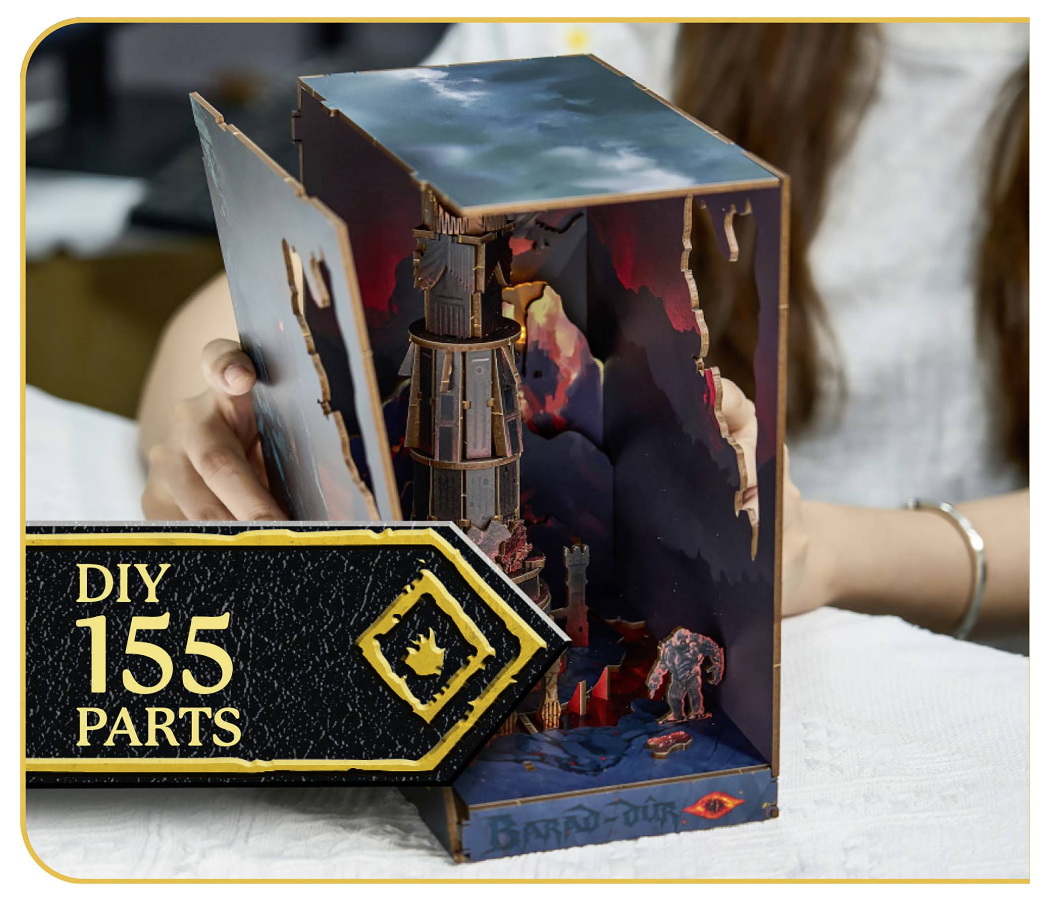 Osoba montująca model 3D DIY z tekstem 'DIY 155 Części' na wewnętrznej części. Model przedstawia wieżę i scenerię krajobrazu.