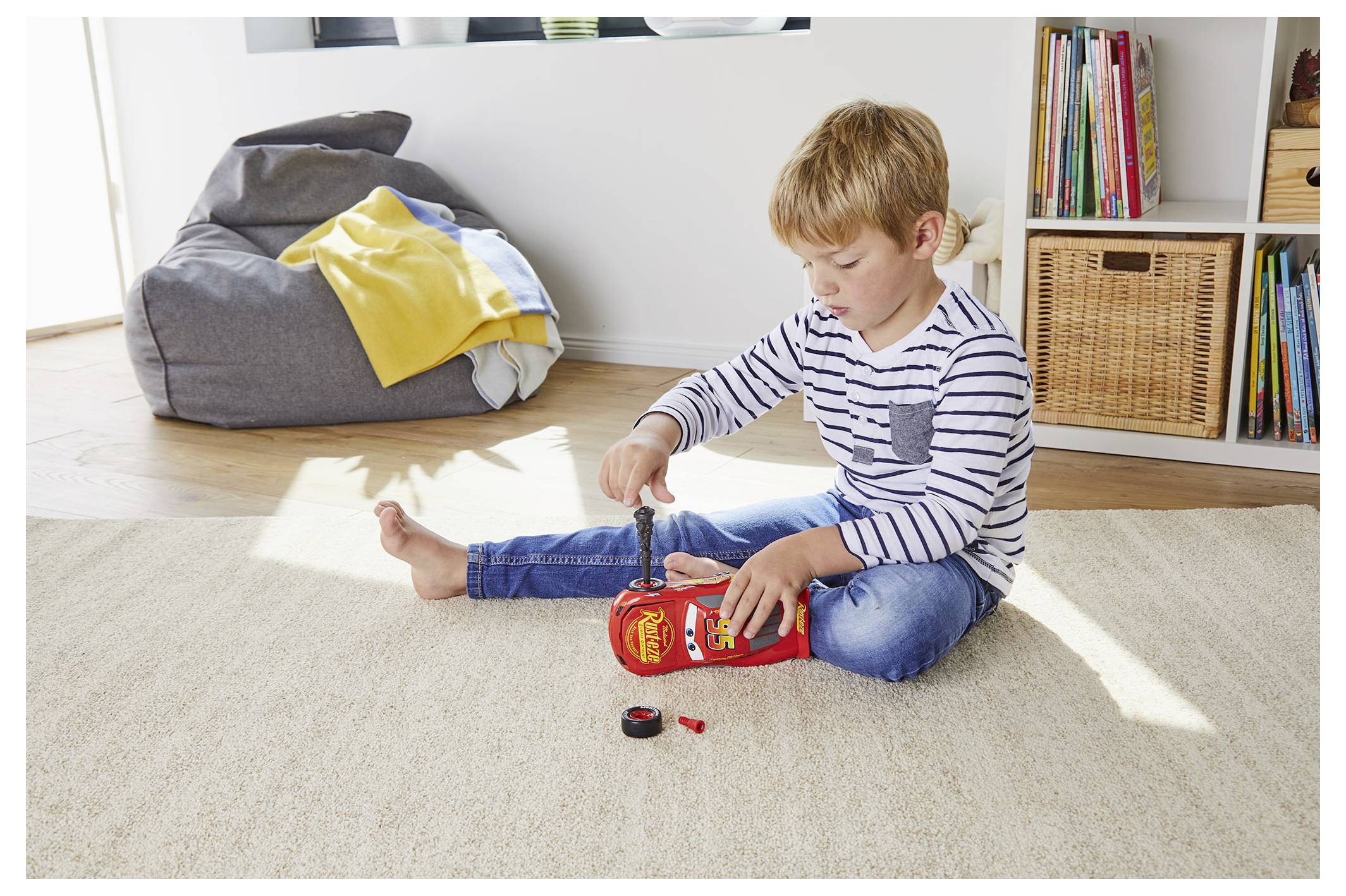 Małe dziecko siedzi na dywanie, bawiąc się miniaturowym samochodzikiem w dobrze oświetlonym pokoju, z workiem sako i regałem na książki w tle.